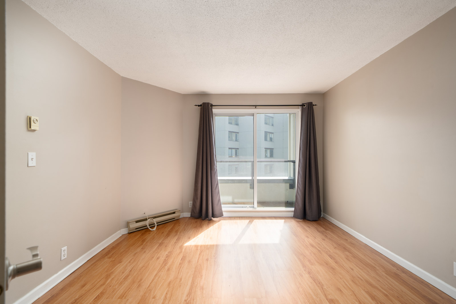 Empty room with light wood flooring, beige walls, a large window with gray curtains, and a small balcony visible outside.