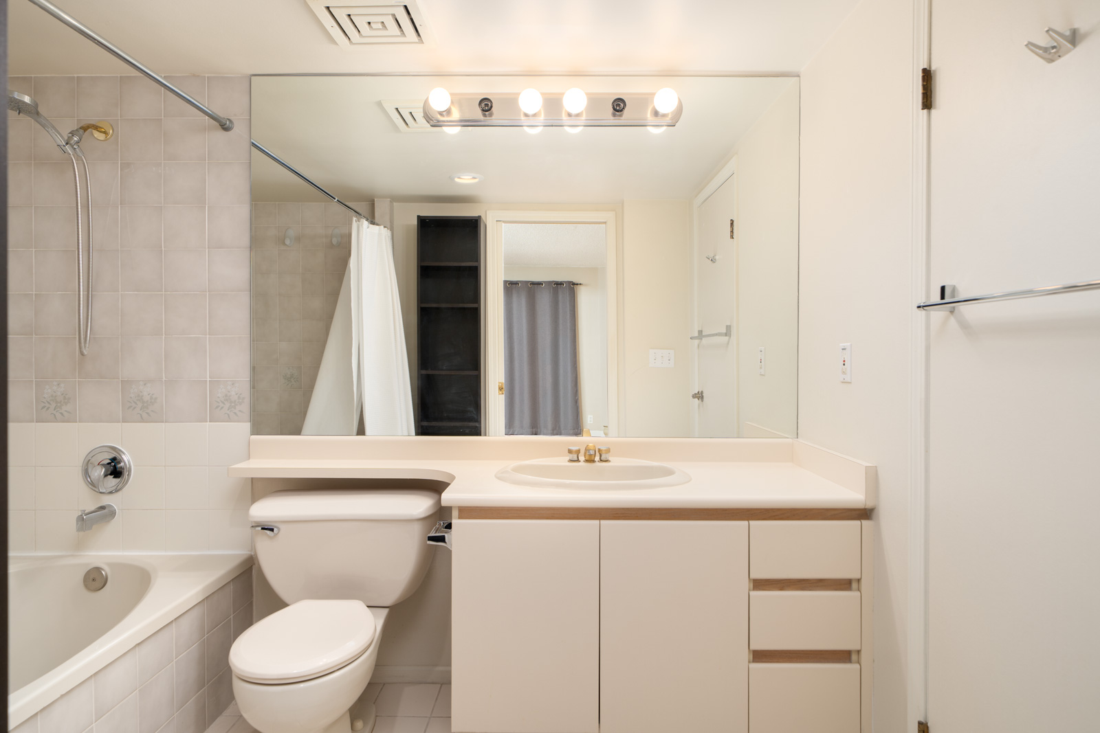 A clean, modern bathroom with a bathtub, toilet, white vanity, large mirror, and overhead lights. A towel bar and shelving are also visible.