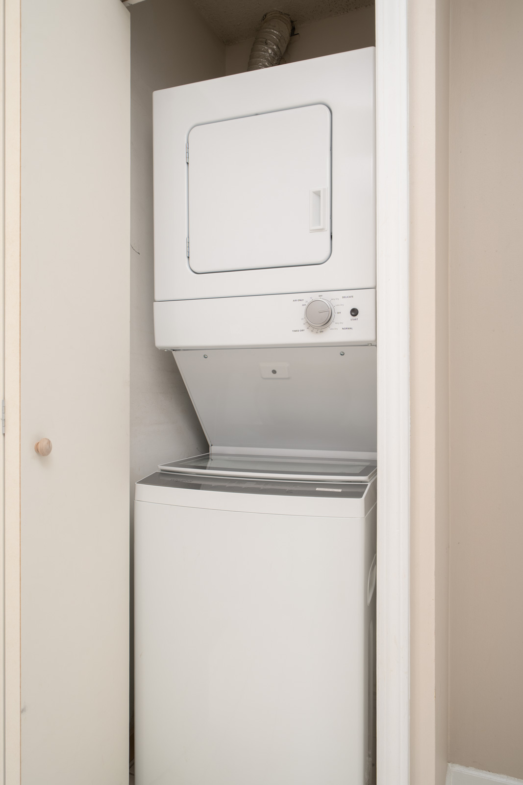 A white stacked washer and dryer unit is installed in a small closet with the dryer door closed and the washer lid open.