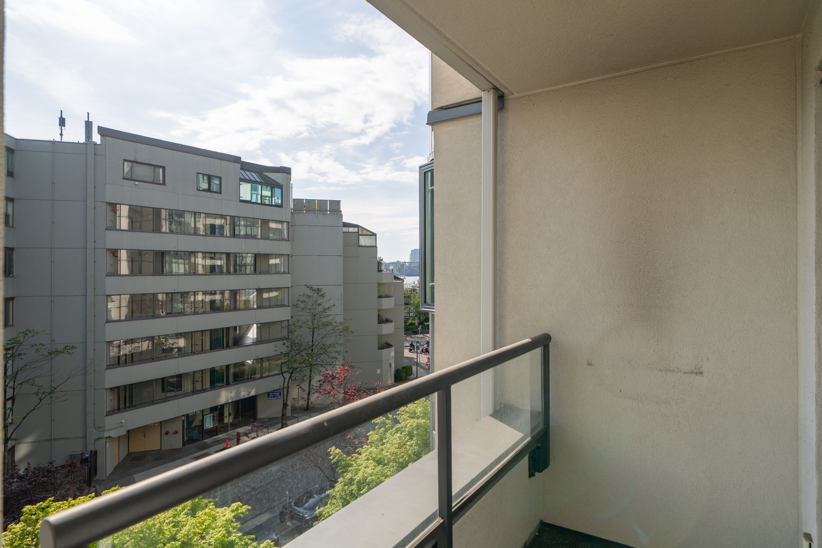 View from a small balcony with a glass railing, overlooking modern apartment buildings and a street below on a sunny day.