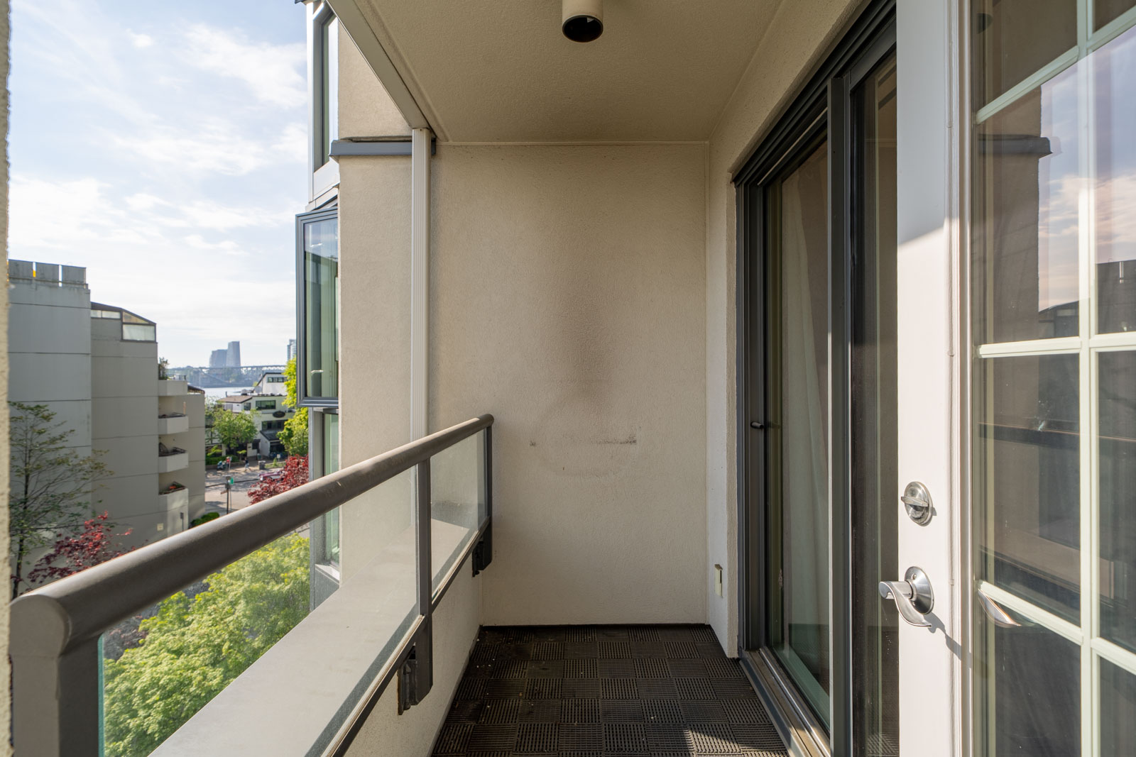 A small, empty apartment balcony with glass railing and sliding glass doors, overlooking nearby buildings and trees on a sunny day.