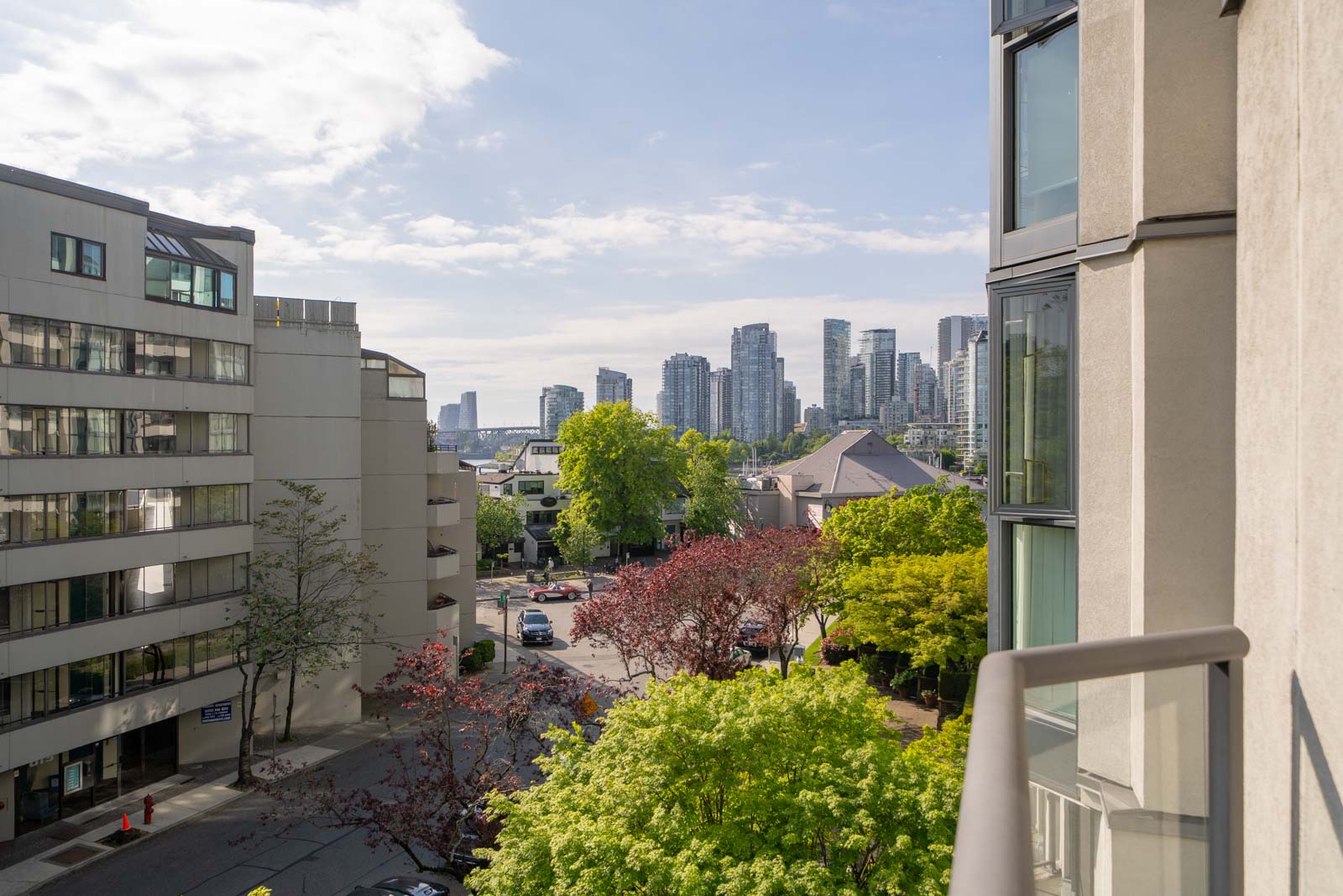 Urban street view with modern apartment buildings, green trees, and a city skyline in the background under a partly cloudy sky.