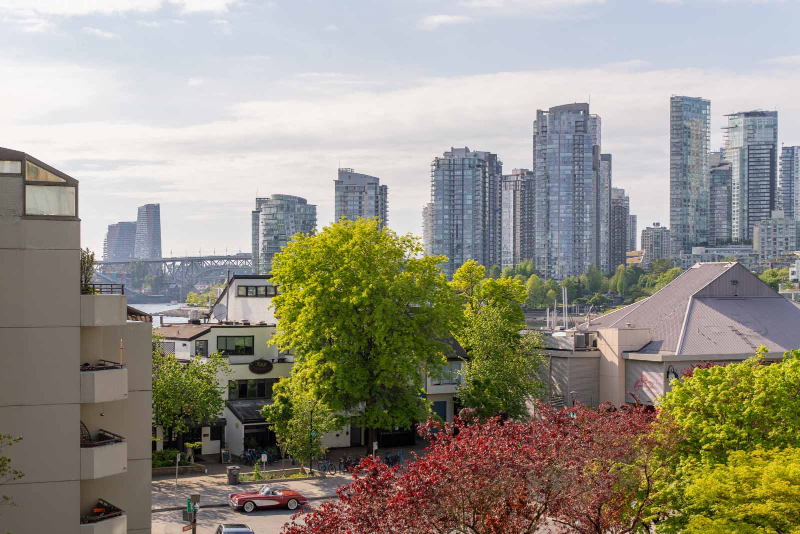 Cityscape view with modern high-rise buildings, trees with green and red foliage, and a few low-rise structures in the foreground under a partly cloudy sky.
