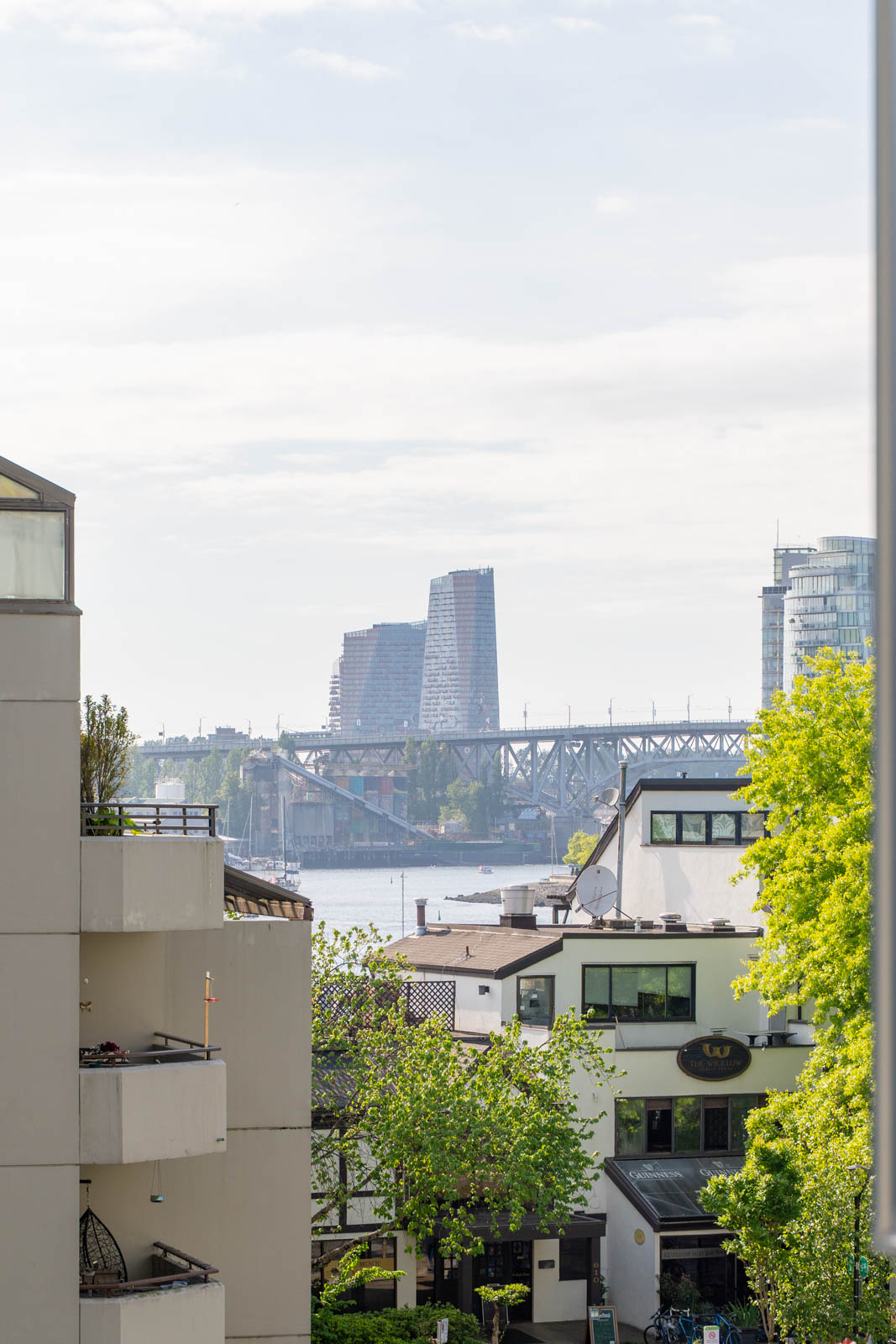 View of modern buildings and a bridge over a body of water, with residential buildings and green trees in the foreground under a partly cloudy sky.