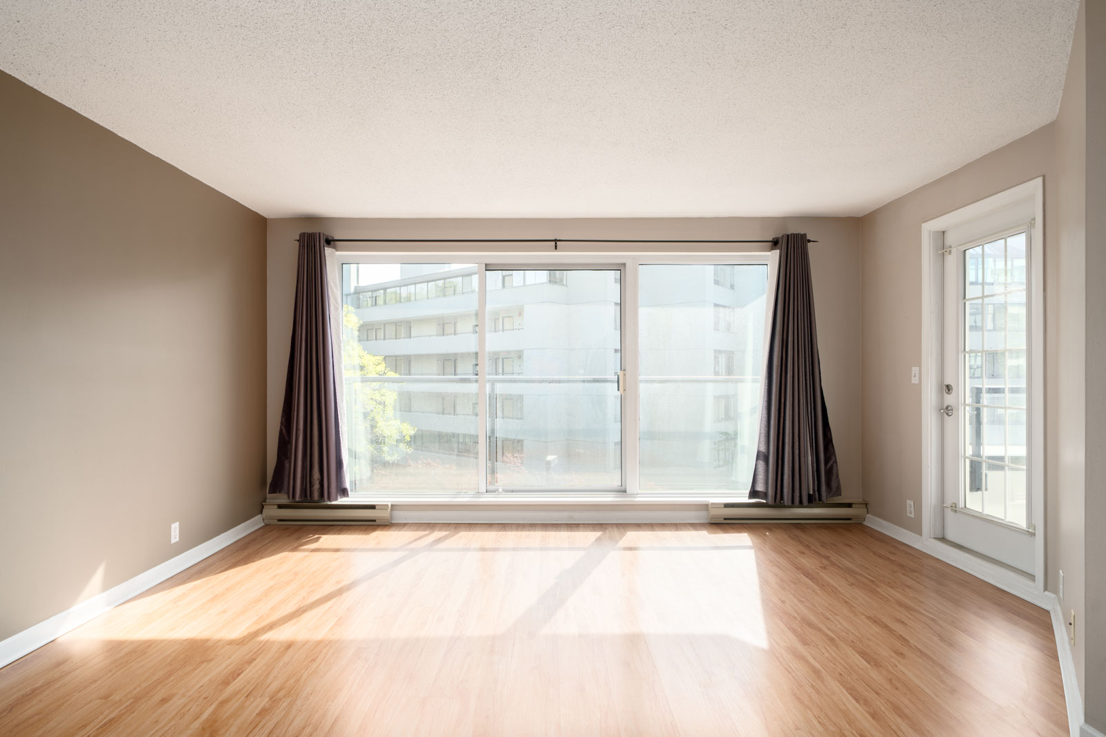 Empty living room with light wood flooring, beige walls, large window and glass door leading to a balcony, with natural light streaming in.