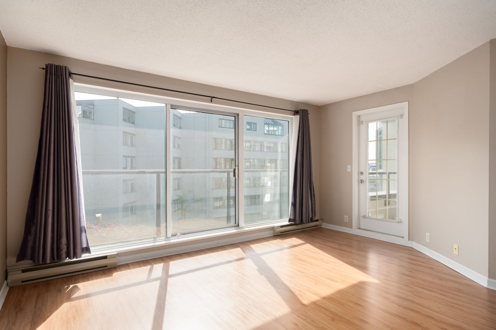 Unfurnished room with wood flooring, large window, glass door, and dark curtains, overlooking apartment buildings in daylight.