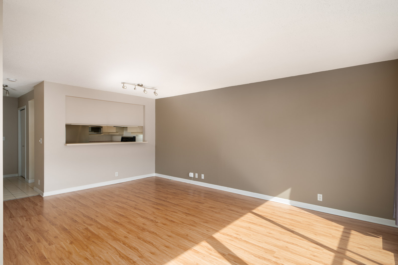 Unfurnished living room with wood flooring, tan walls, large window with sunlight, and an open pass-through to the kitchen area.