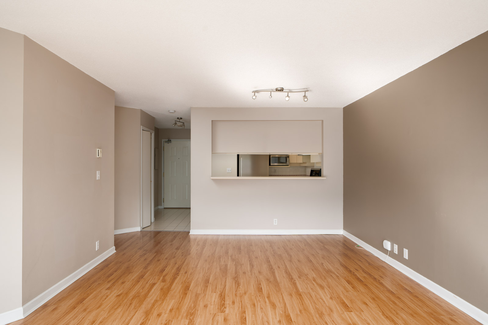 Unfurnished living room with light wood flooring, beige walls, a pass-through window to the kitchen, and a ceiling light fixture. Doorways to other rooms are visible in the background.