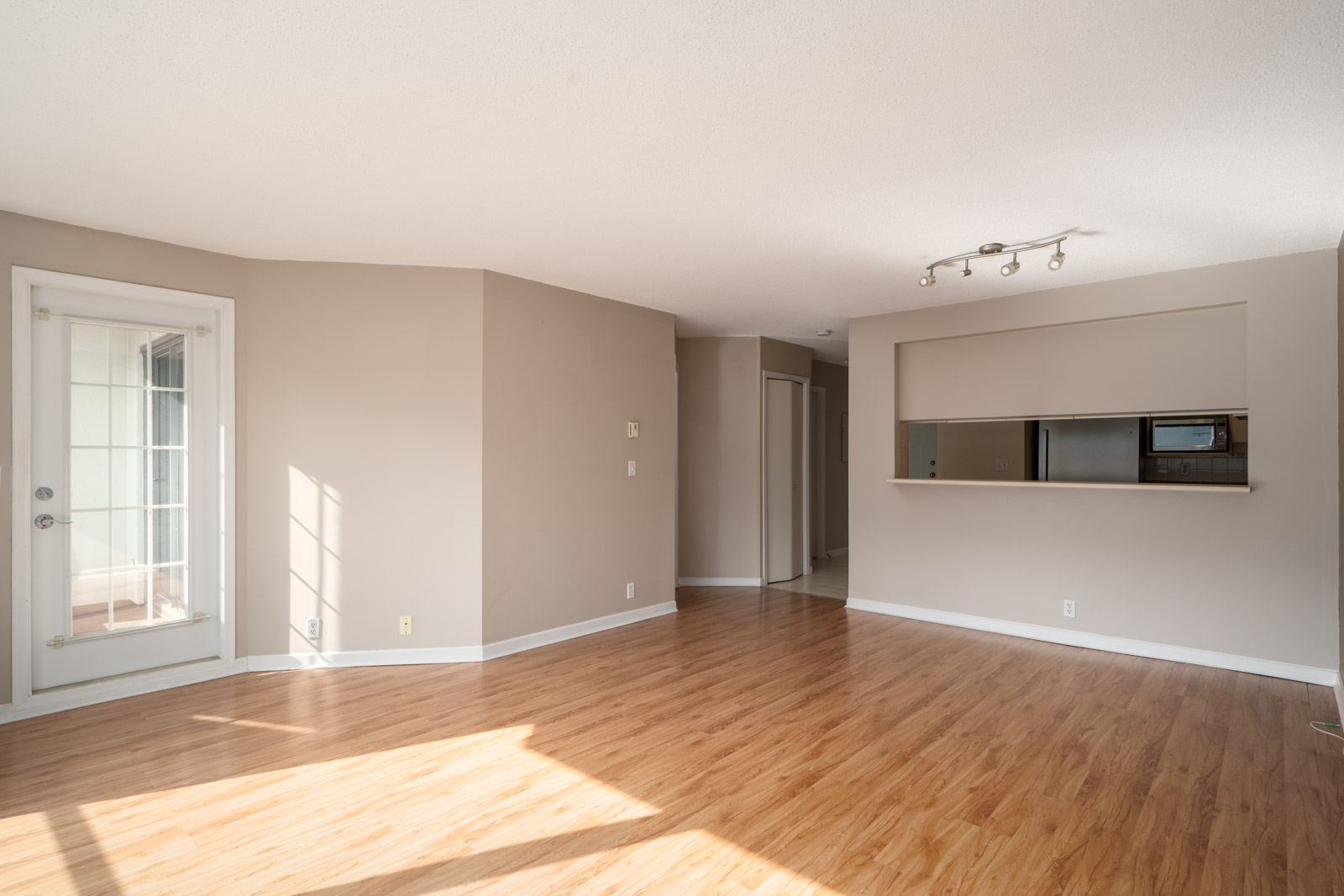 Empty living room with light wood flooring, beige walls, a glass door to the left, and an open pass-through window to the kitchen on the right. Ceiling track lights are visible.