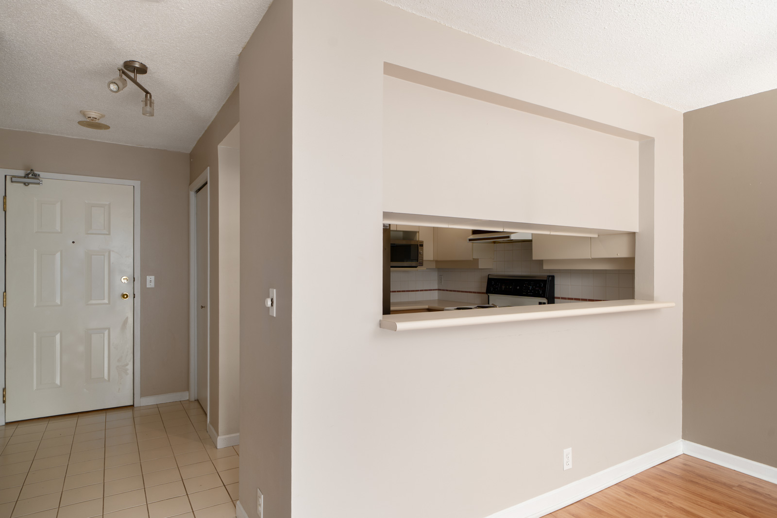 Apartment interior showing an entryway with a white door, hallway, and a passthrough window to a kitchen with beige walls and light flooring.