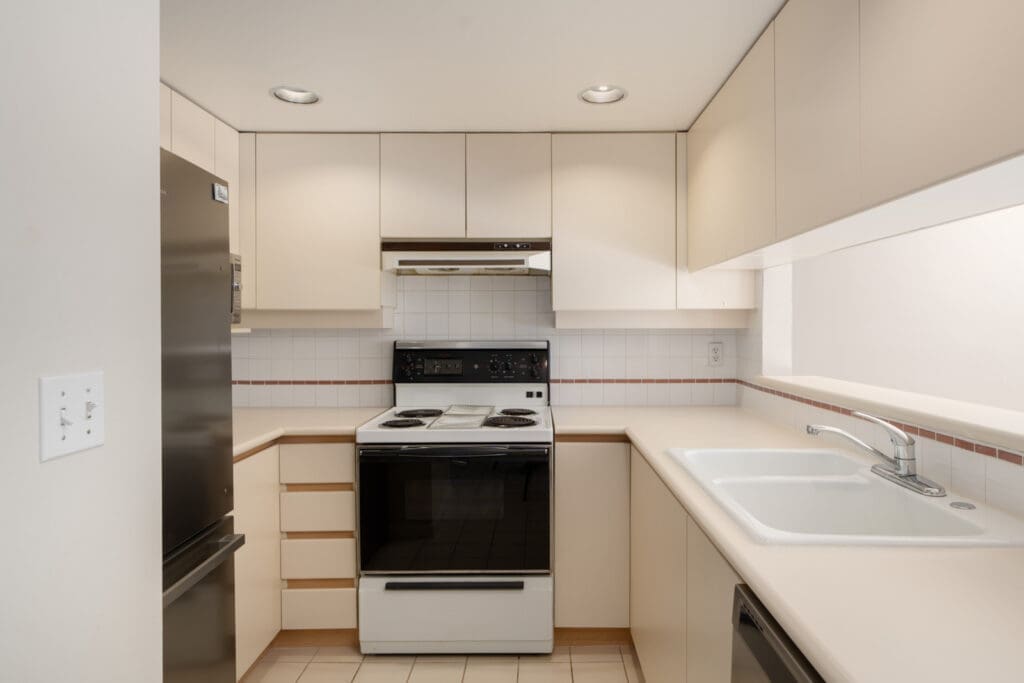 Small kitchen with beige cabinets, white countertop, black and white stove, refrigerator, sink, and tiled backsplash. Ceiling lights and some drawer handles are missing.
