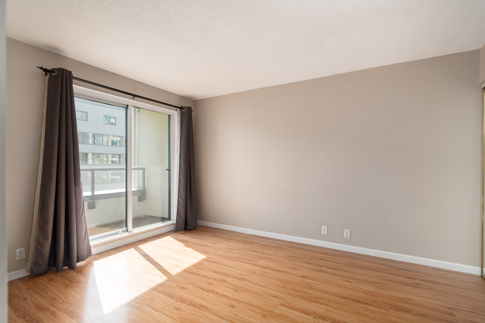 Empty room with light wood flooring, beige walls, a large window with dark curtains, and a sliding glass door leading to a balcony. Sunlight shines through the window.