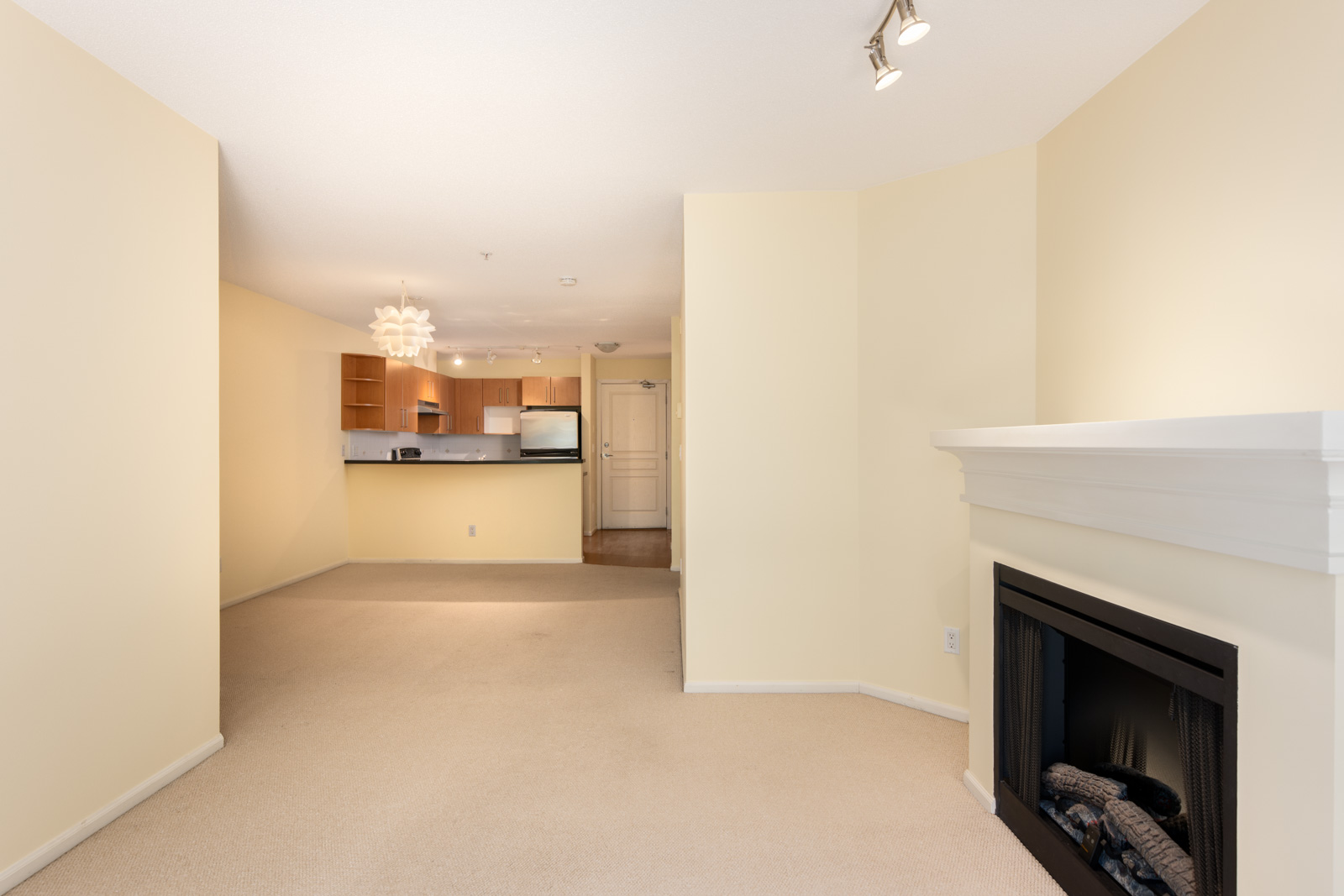 A living room with beige carpet and walls, a white fireplace on the right, and an open view into a kitchen with wooden cabinets and stainless steel appliances.