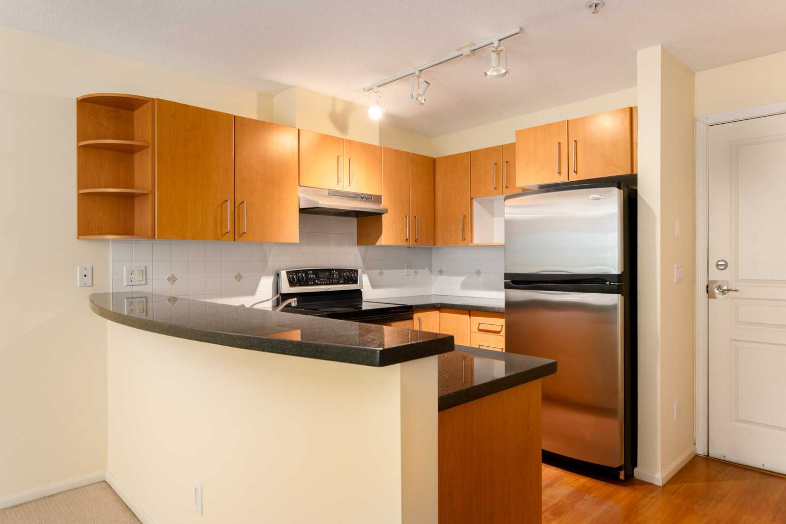 Modern kitchen with light wood cabinets, stainless steel appliances, black countertops, white tile backsplash, and overhead track lighting.