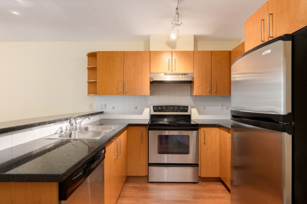 Modern kitchen with stainless steel appliances, black countertops, wooden cabinets, a double sink, and track lighting above.