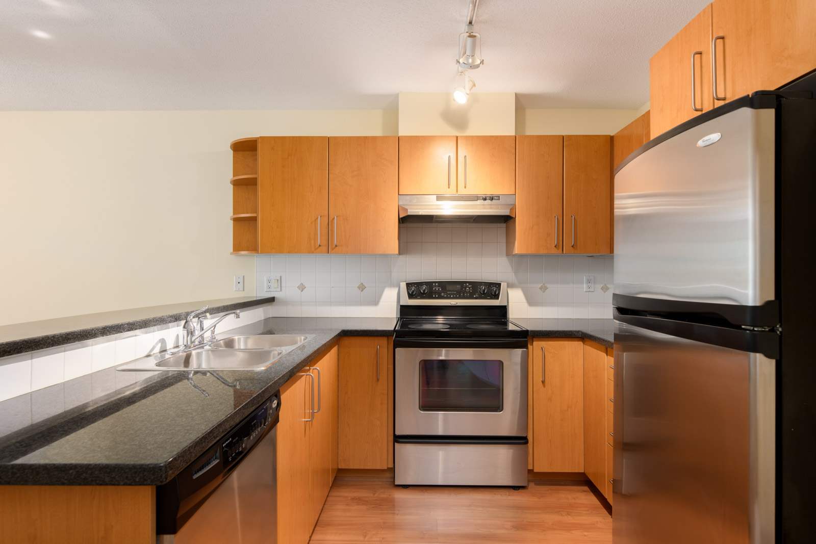 Modern kitchen with stainless steel appliances, black countertops, wooden cabinets, a double sink, and track lighting above.