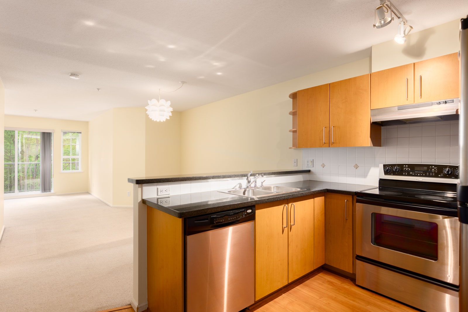 Modern kitchen with wood cabinets, stainless steel appliances, and black countertops, open to a bright living area with large windows and access to a balcony or patio.