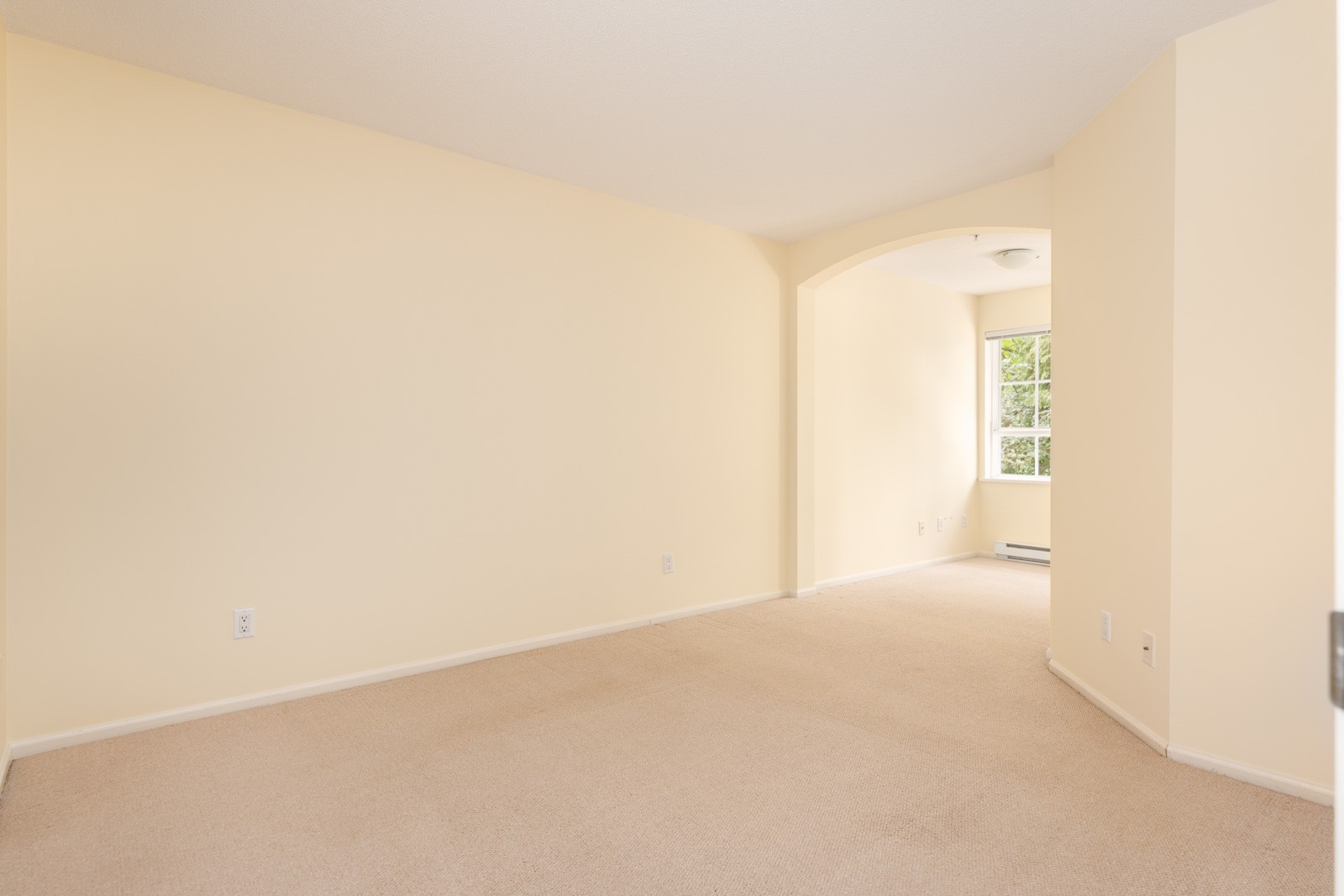 An empty room with beige carpet and light yellow walls, featuring an arched doorway leading to a window with natural light.