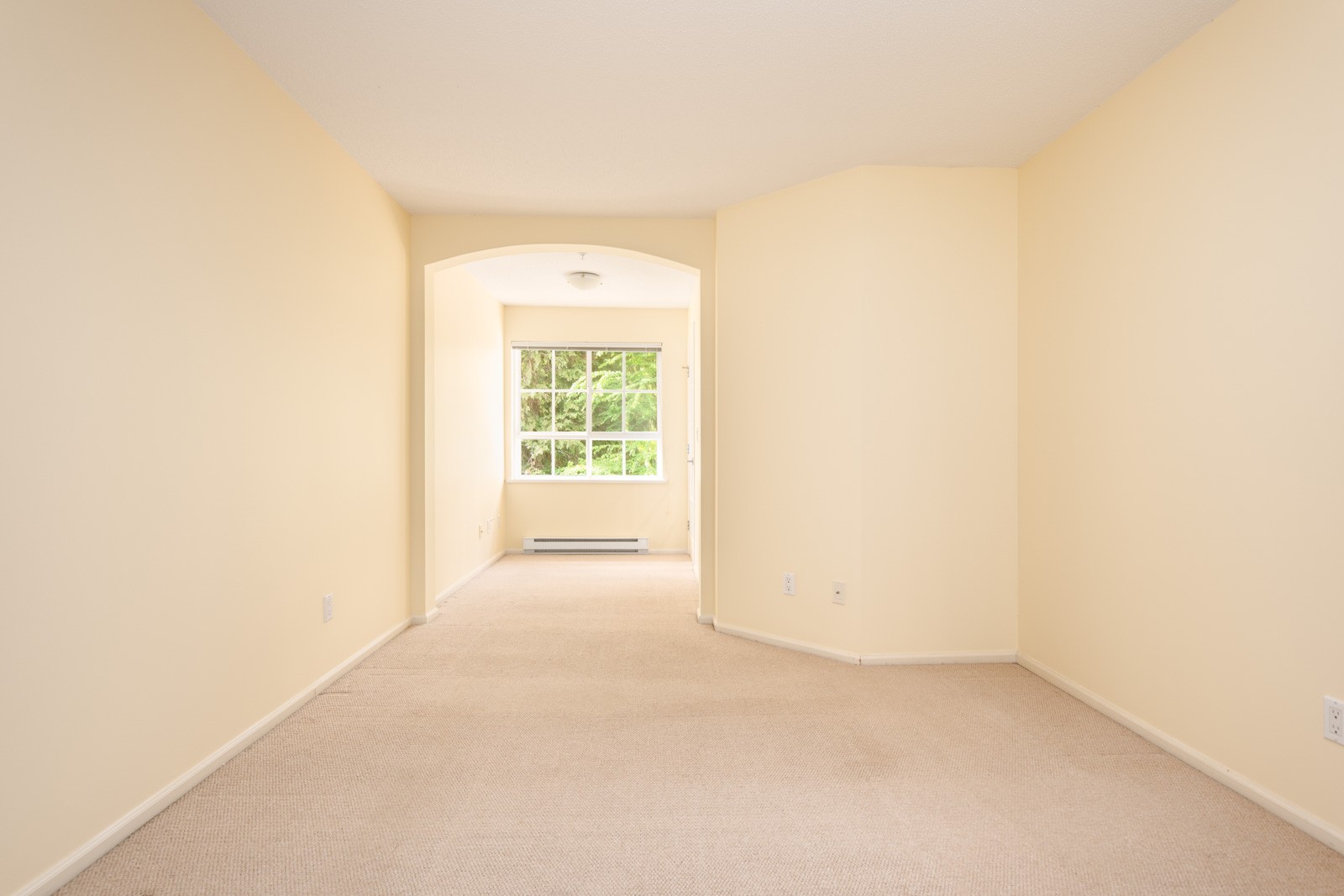 Empty, carpeted room with cream-colored walls, a large window at the far end letting in natural light, and an arched entryway leading to the window area.