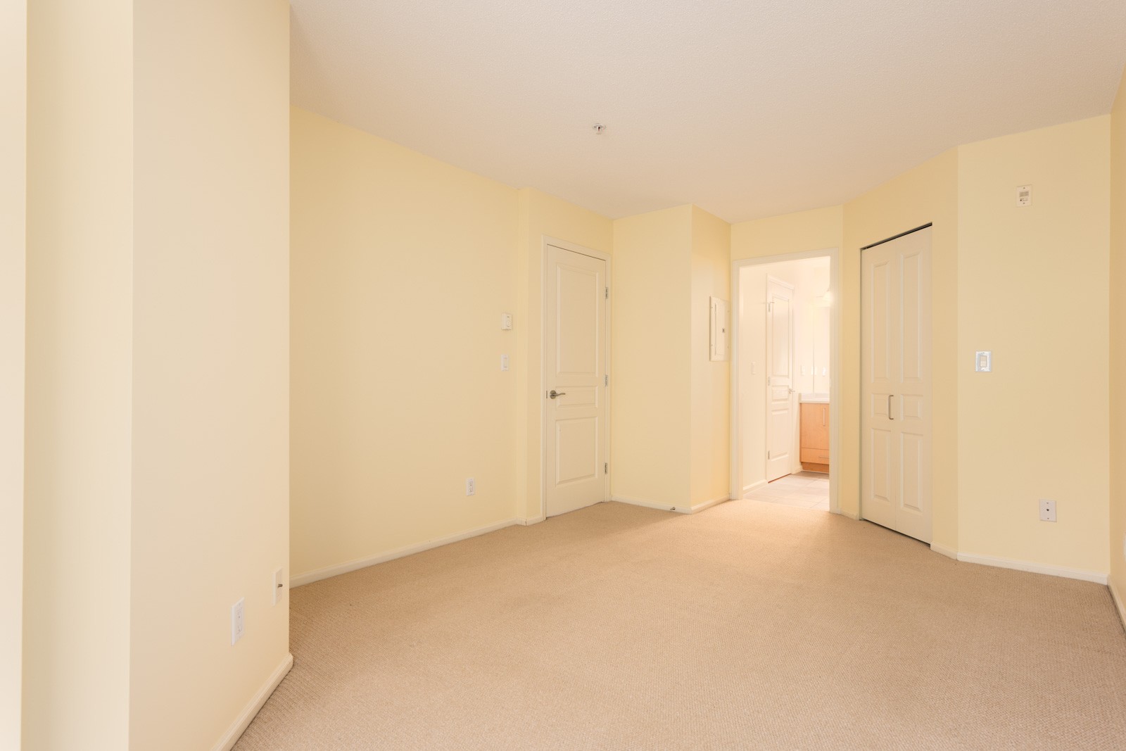 Empty beige carpeted room with light yellow walls, several doors including a closet, and an open doorway leading to a well-lit bathroom.