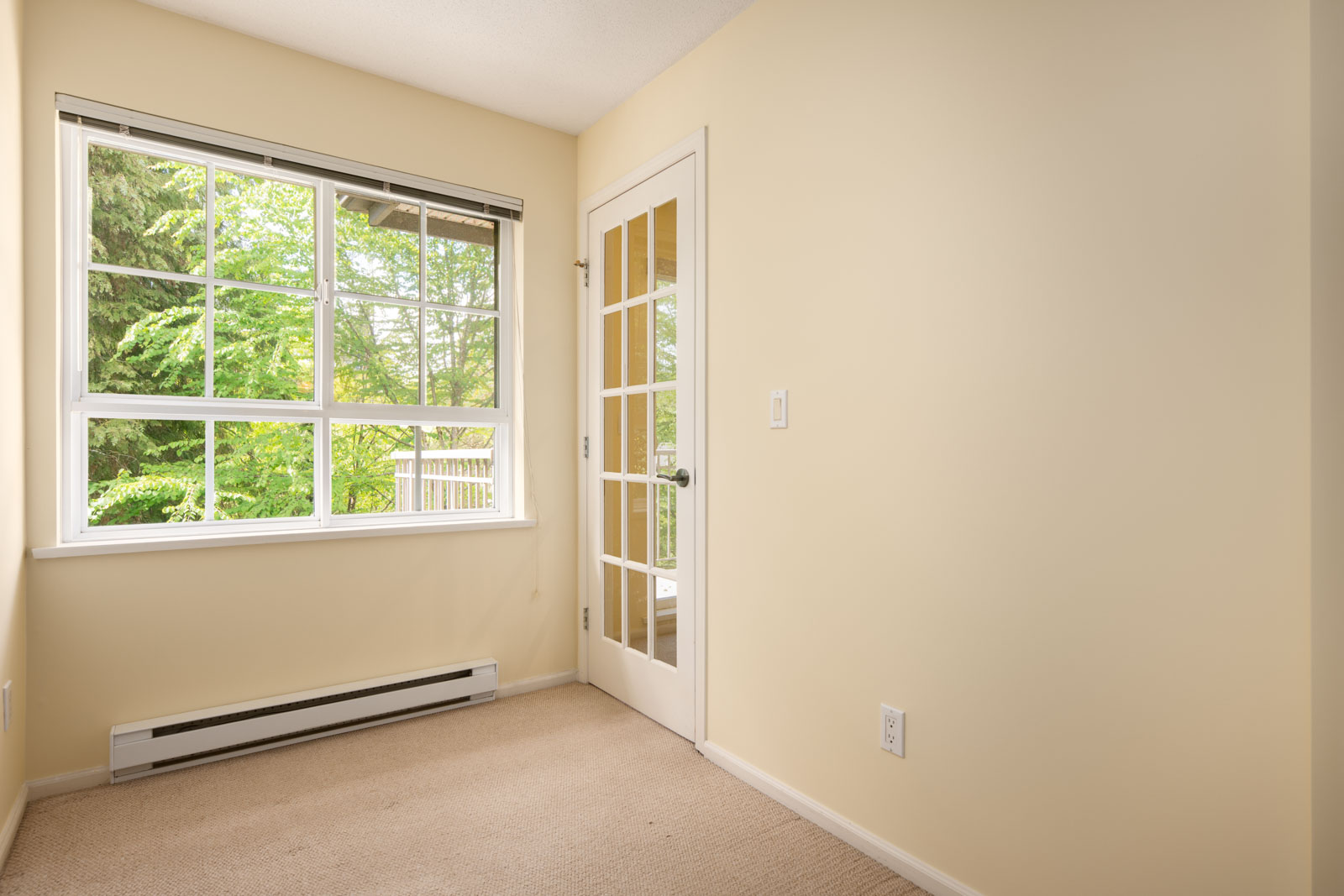 Small empty room with beige carpet, pale yellow walls, a large window with outdoor view, and a glass-panel door.