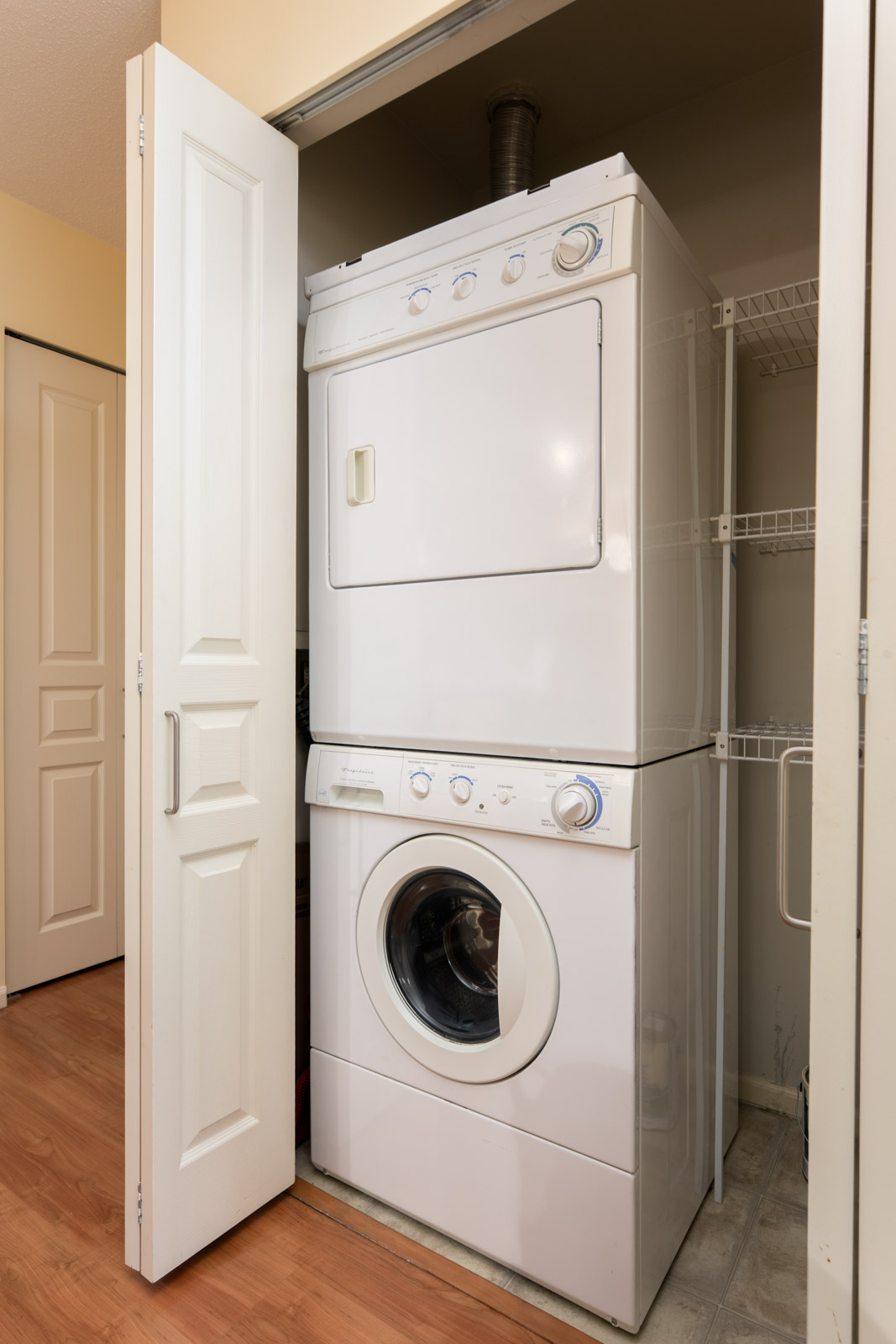 A stacked washer and dryer unit is installed in a small laundry closet with white folding doors and wire shelving on the side.
