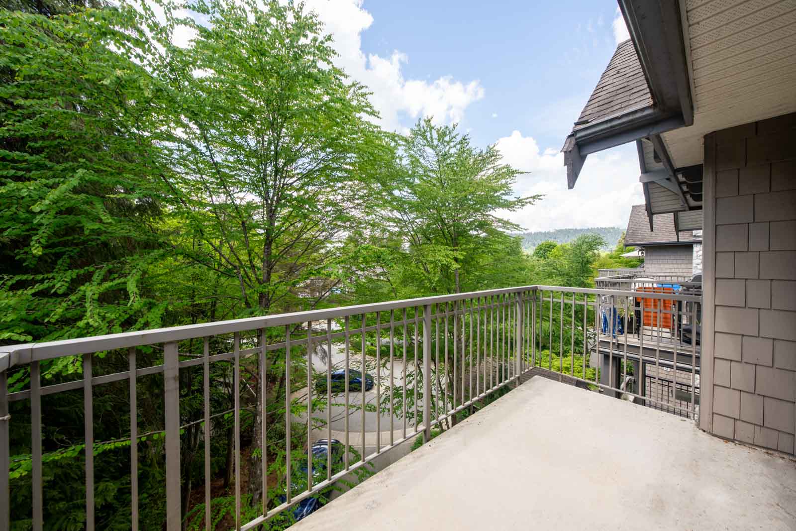 Second-floor balcony with metal railing overlooking trees, a parking area, and neighboring buildings under a partly cloudy sky.