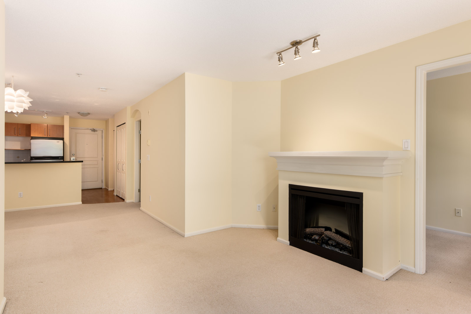 Unfurnished living room with beige walls and carpet, a white fireplace, and view into the kitchen with stainless steel appliances.