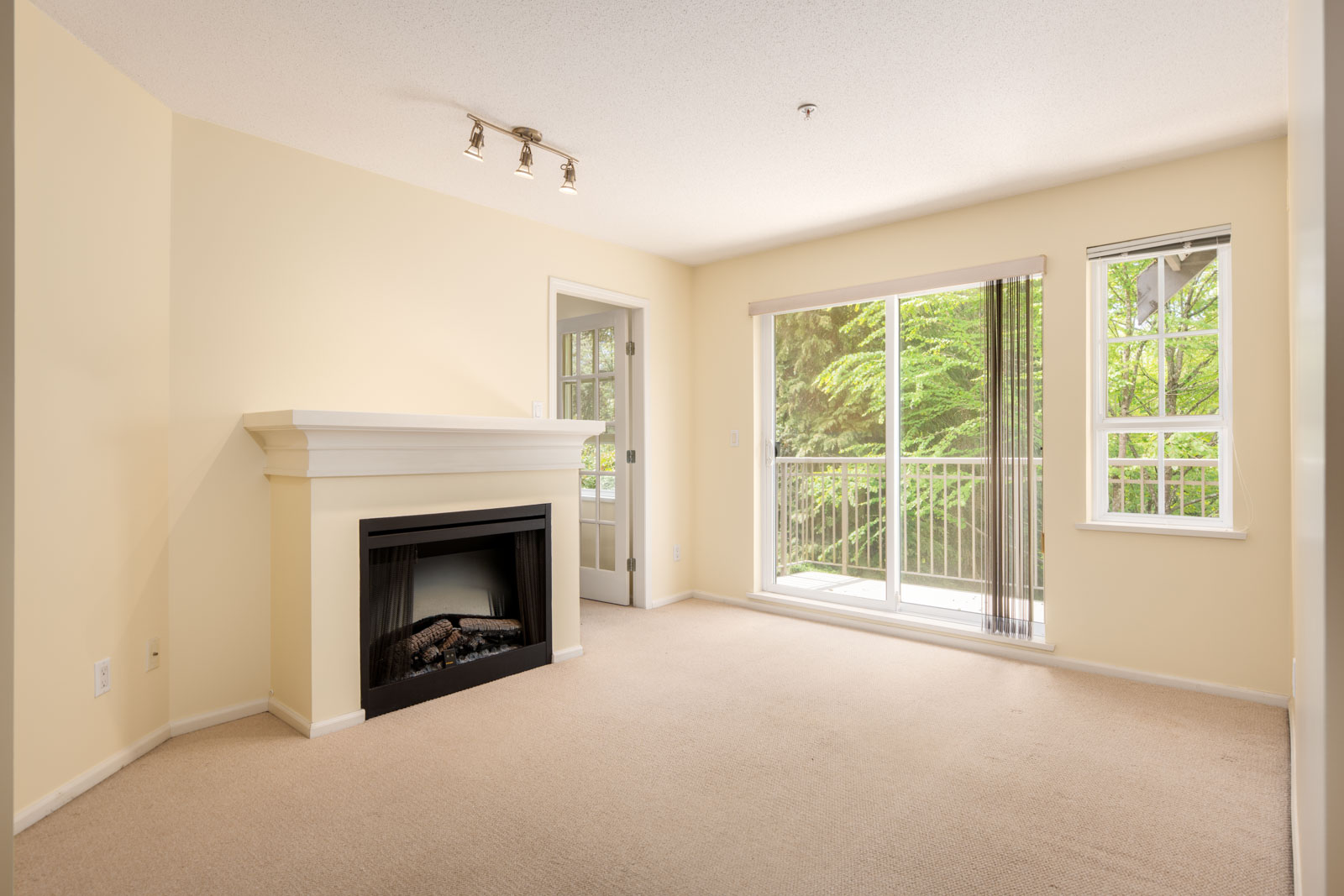 Unfurnished living room with beige carpet, gas fireplace, cream walls, sliding glass door, and windows overlooking a balcony with greenery outside.