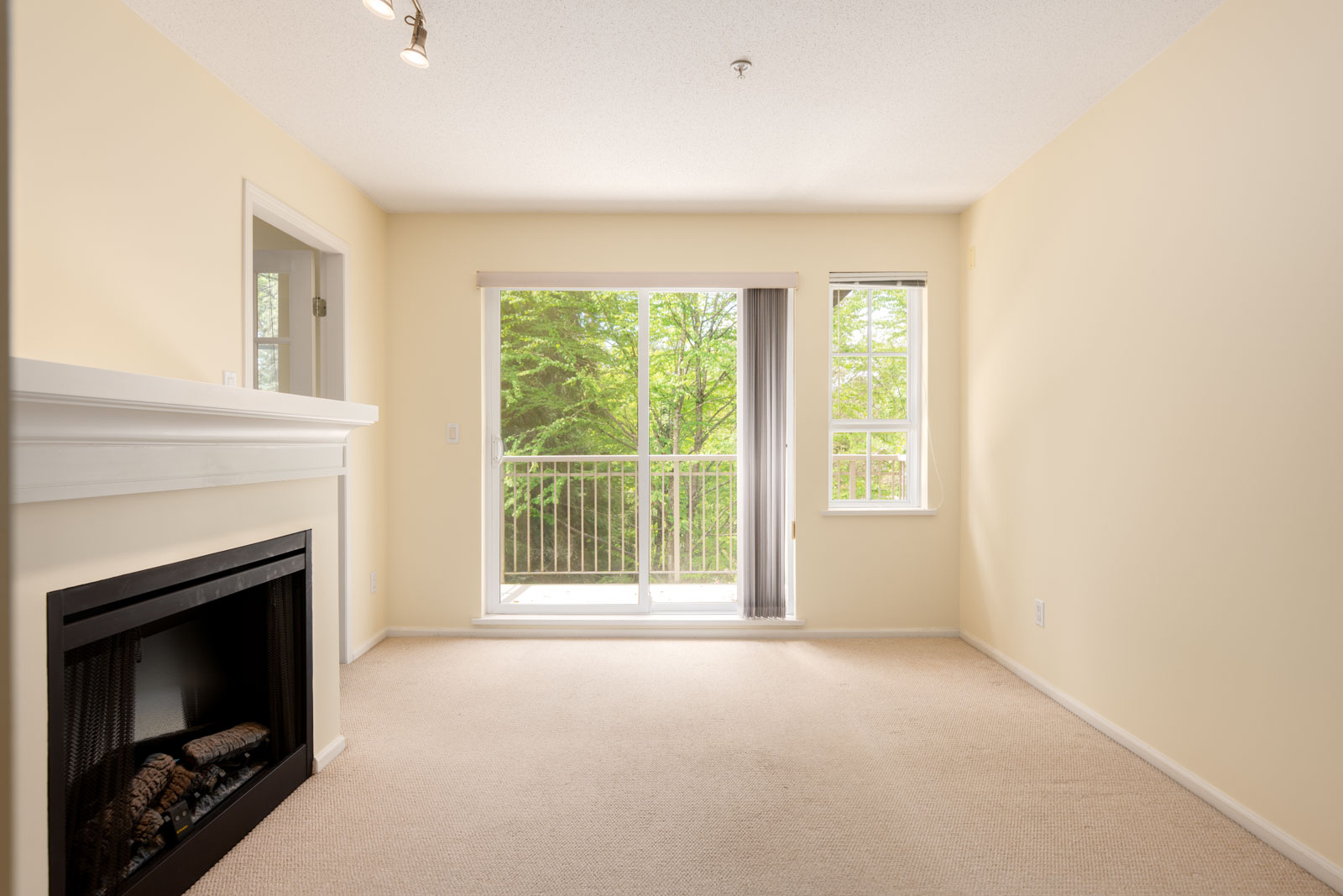 A bright, empty living room with beige carpet, a white fireplace, and large sliding glass doors leading to a balcony with a view of green trees.