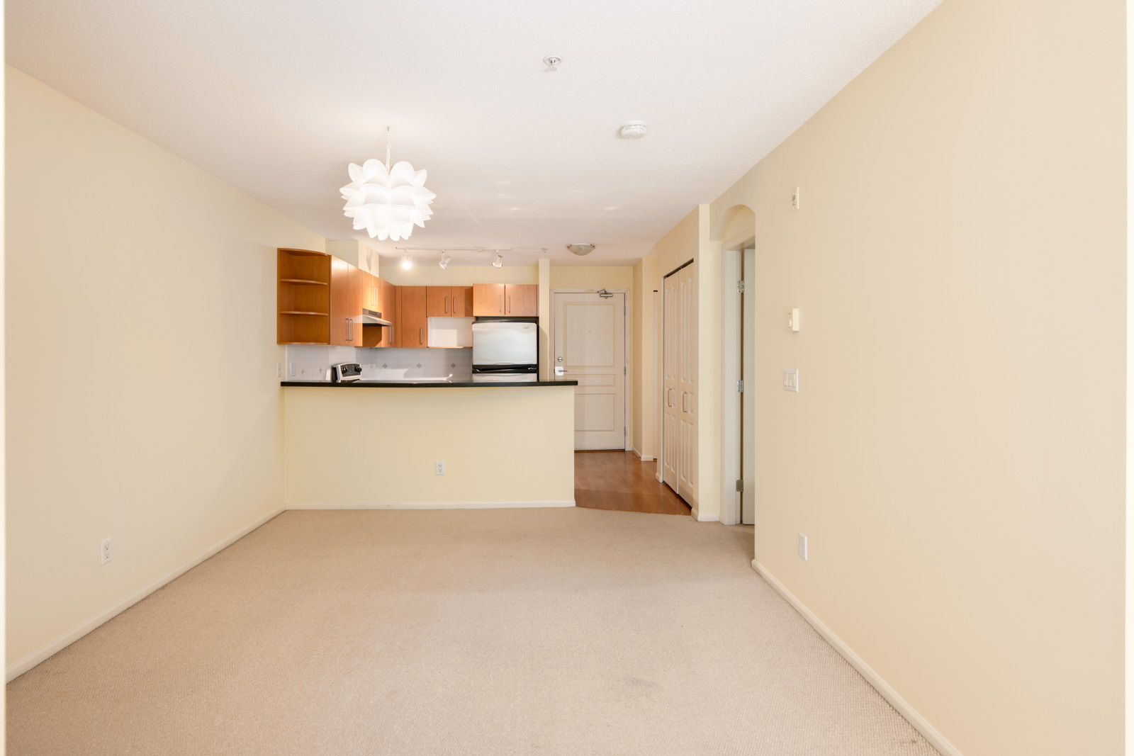 Unfurnished apartment interior with beige walls and carpet, open kitchen with wood cabinets and stainless steel appliances, and a white pendant light fixture.
