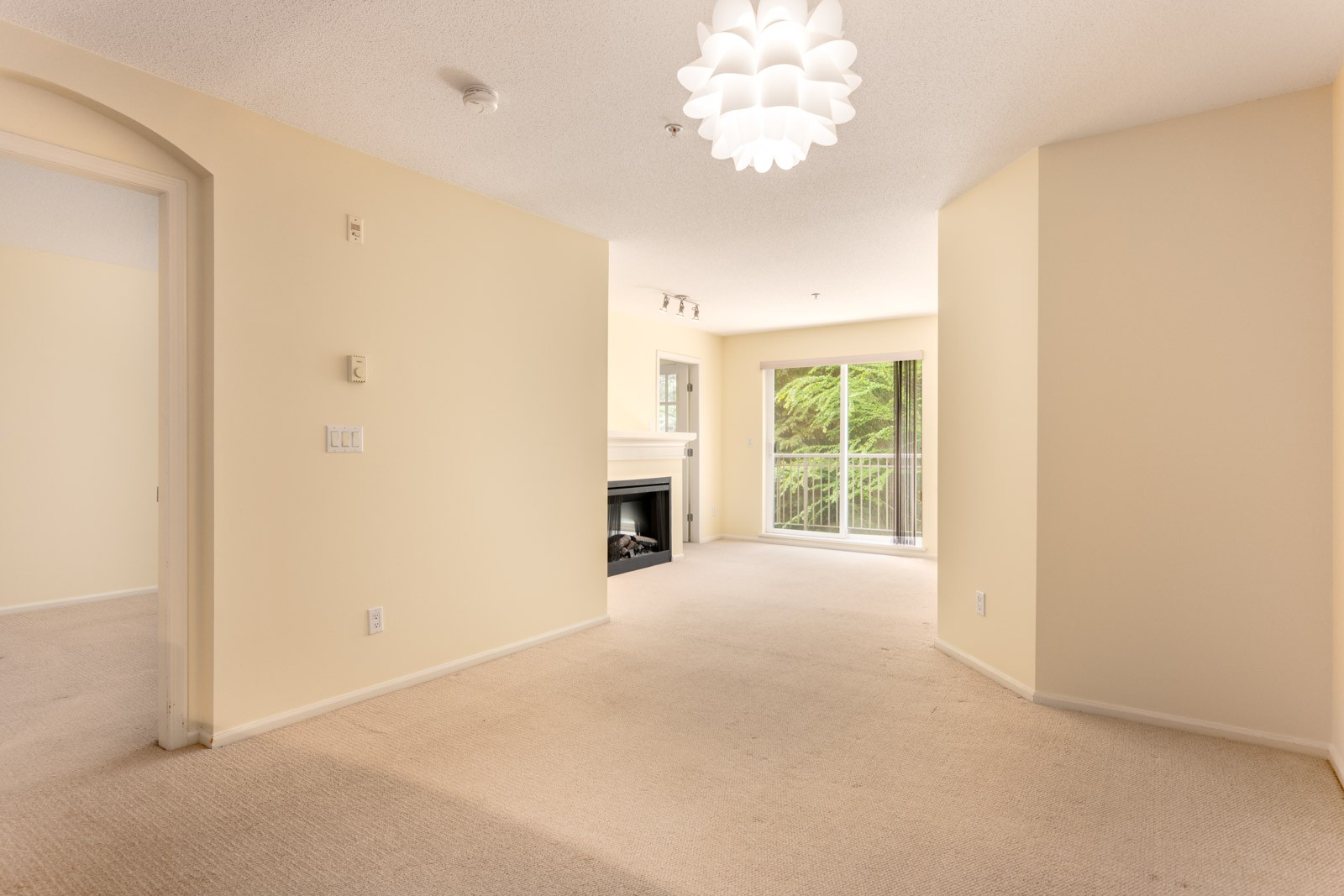 Unfurnished living room with beige carpet, cream walls, a modern ceiling light, fireplace, and sliding glass doors leading to a balcony with greenery outside.