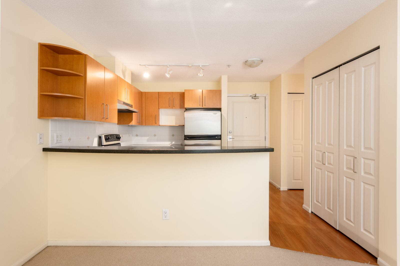 A small modern kitchen with light wood cabinets, stainless steel appliances, white tile backsplash, and a peninsula counter, adjacent to a hallway and double closet doors.