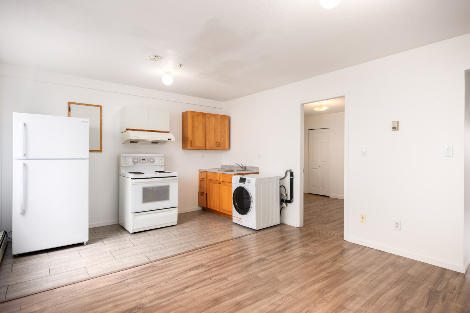 A small kitchen features a white fridge, stove, range hood, washing machine, wooden cabinets, and a sink, with a mix of tile and wood flooring.