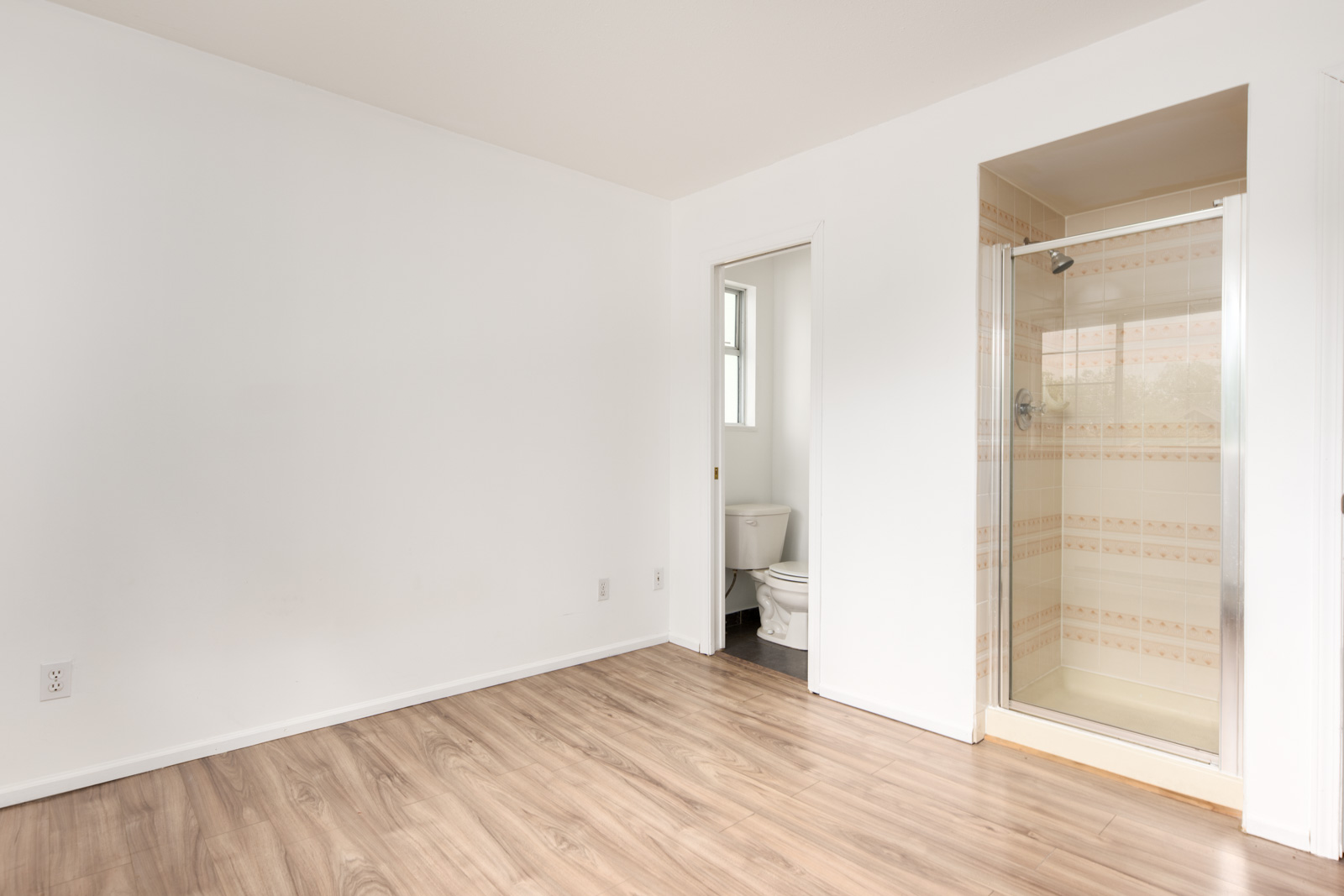 Room with light wood flooring, white walls, an open doorway to a small bathroom with a toilet, and a glass shower enclosure.