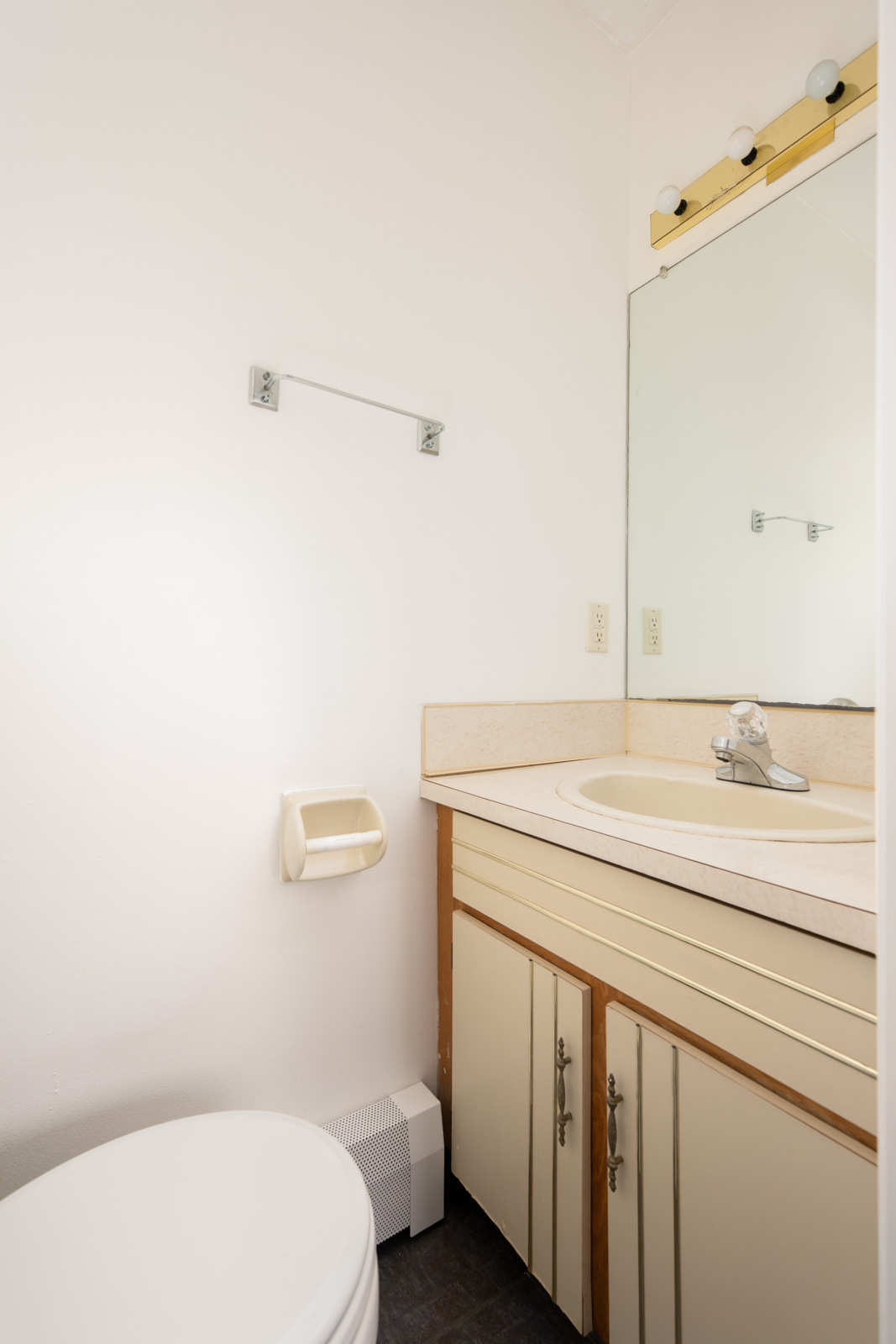 Small bathroom with a white toilet, beige sink countertop, under-sink cabinet, wall mirror, towel rack, and exposed vanity light bulbs above the mirror.