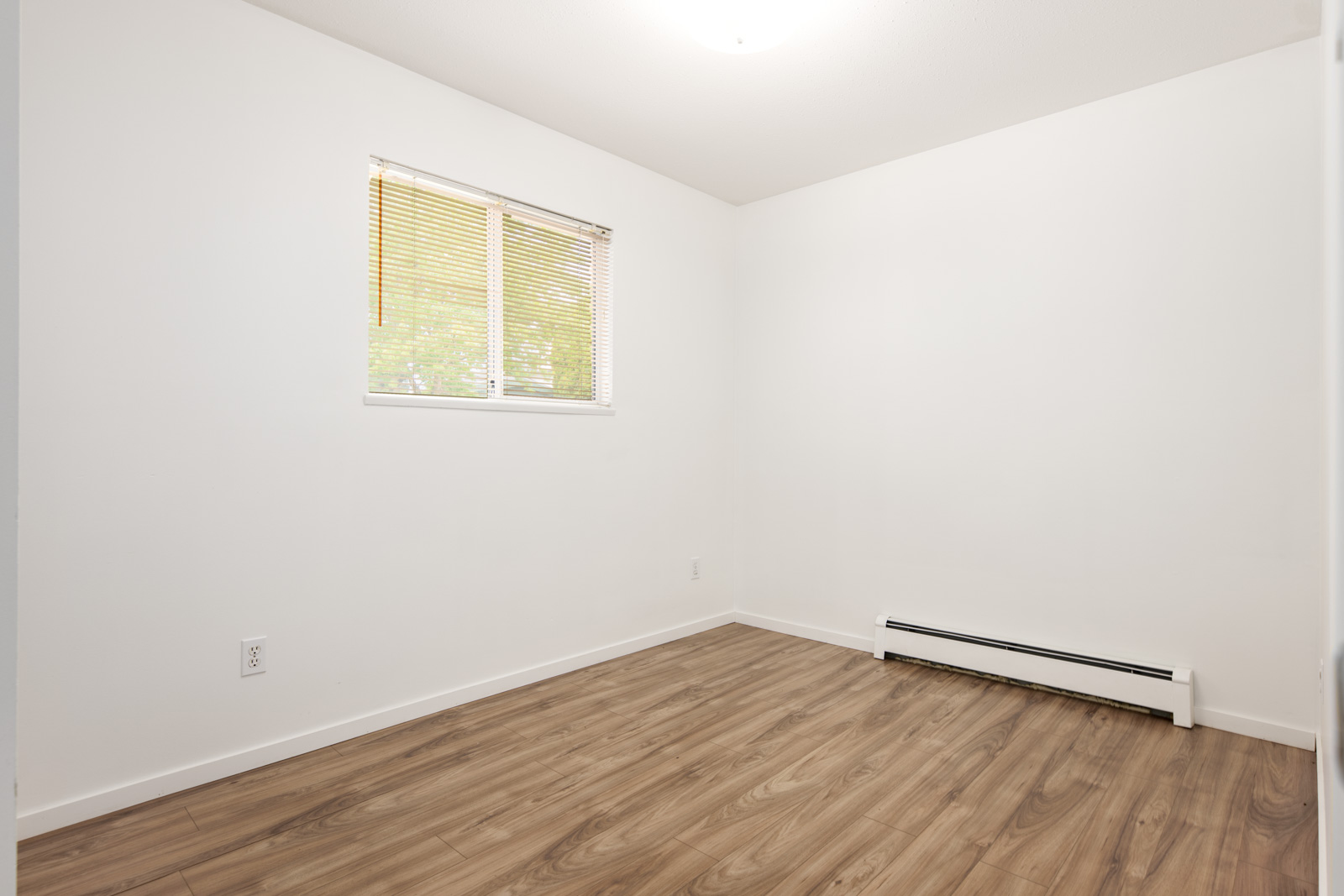 Empty room with white walls, a small window with blinds, wood laminate flooring, and a baseboard heater along one wall.