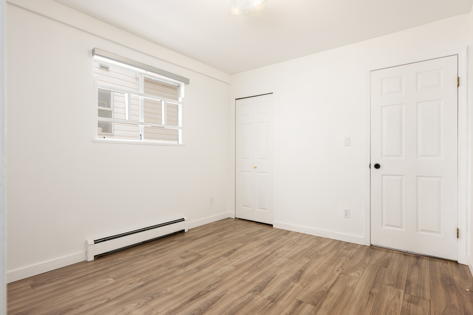 Empty room with white walls, wood-style flooring, a small window with a blind, a baseboard heater, and two closed white doors.