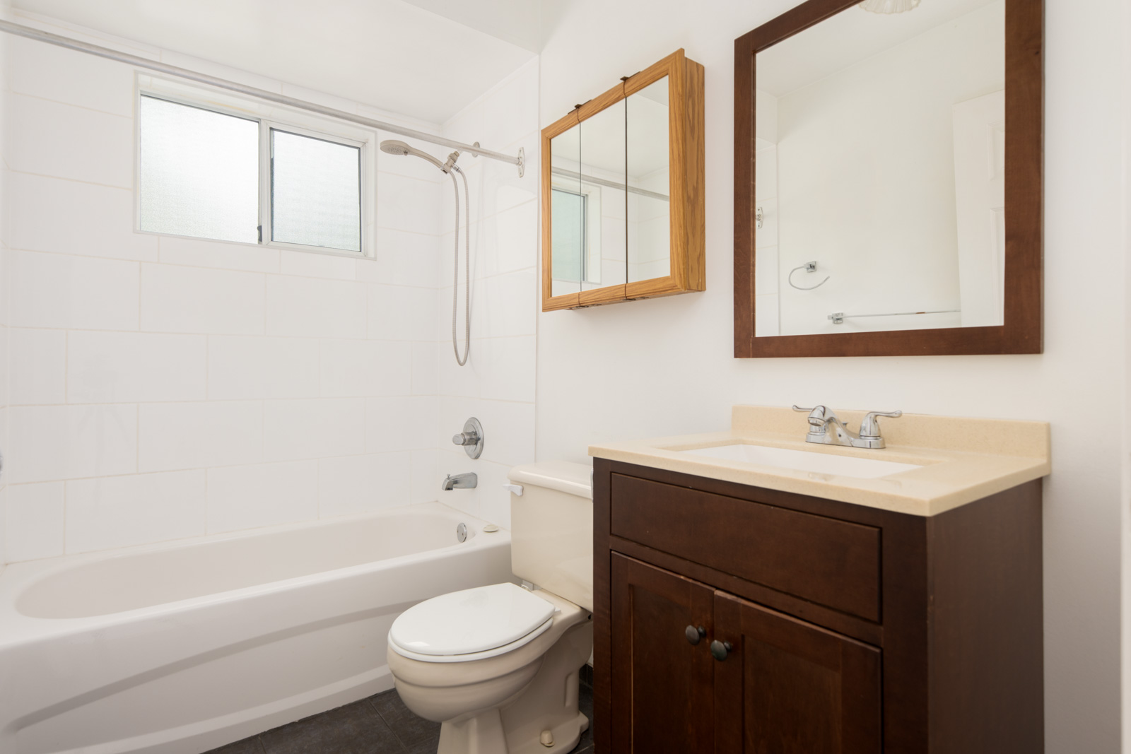 A bathroom with a bathtub and shower, a toilet, a dark wood vanity with sink, a wall mirror, and a window above the tub.