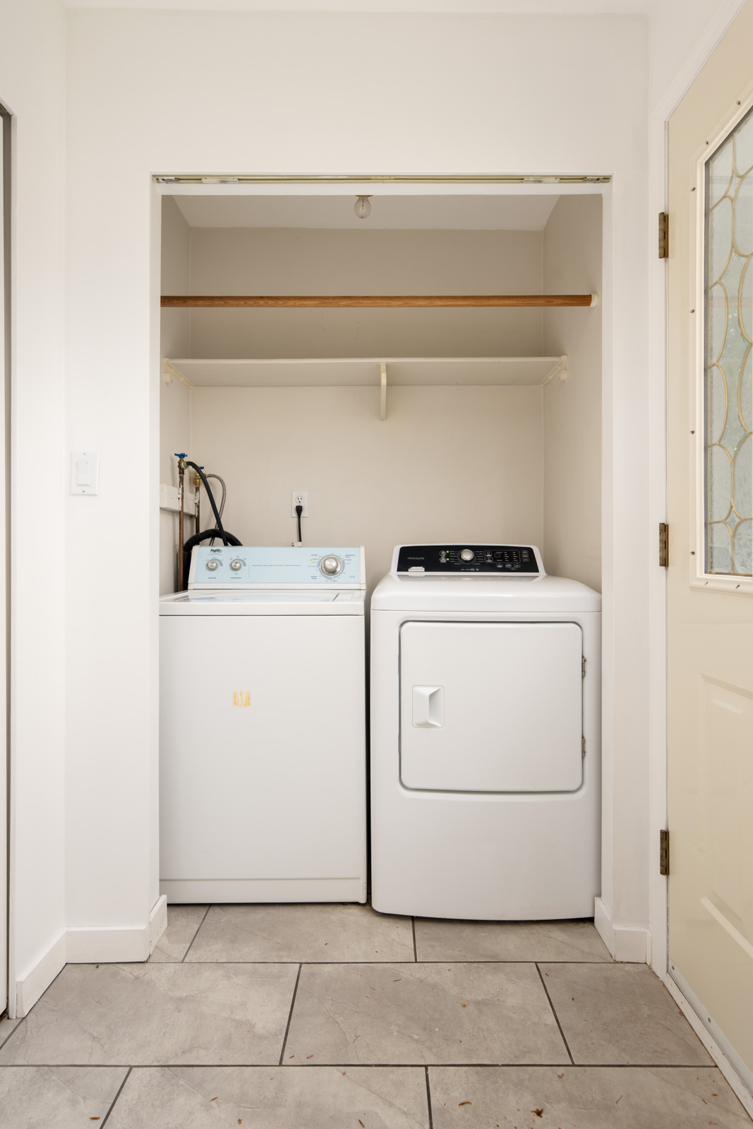 A laundry area with a top-loading washer and a front-loading dryer side by side, under an empty wooden shelf, set against a white wall with a tiled floor.