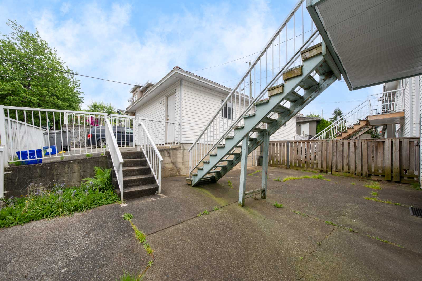 Concrete backyard area with two sets of stairs, one metal and one concrete, leading to a white house and a fenced area, under a partly cloudy sky.