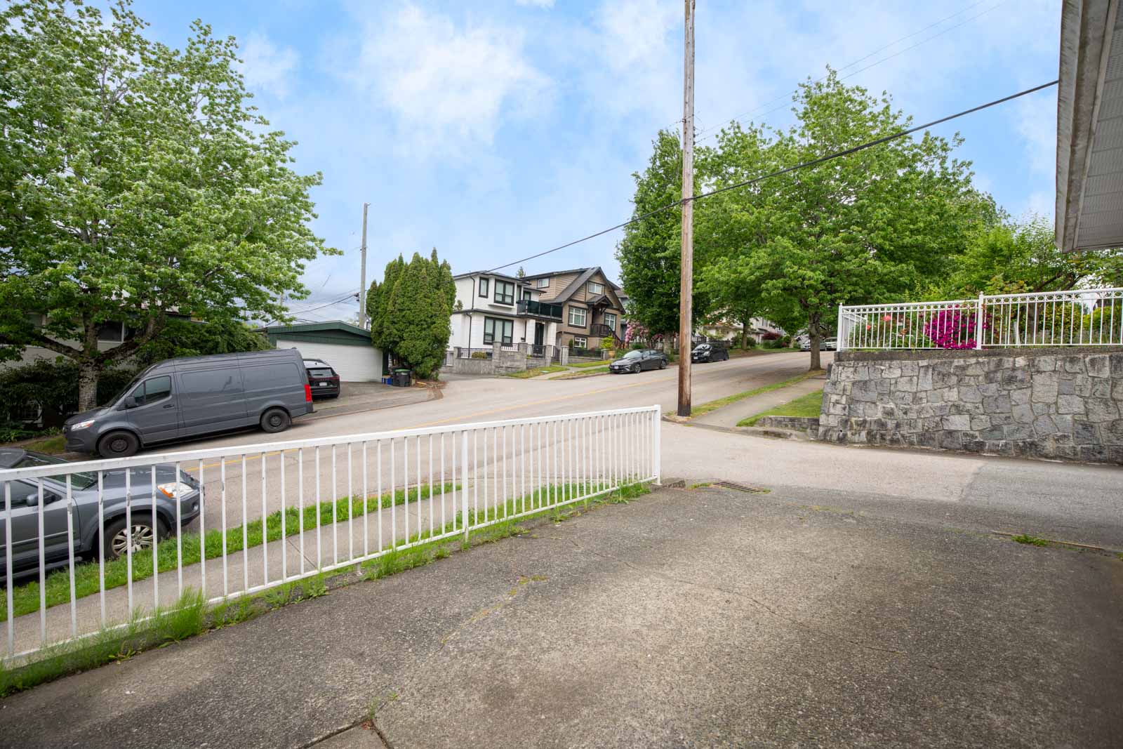 View from a driveway showing a residential street with parked vehicles, houses, trees, a stone wall, and a white metal fence in the foreground.