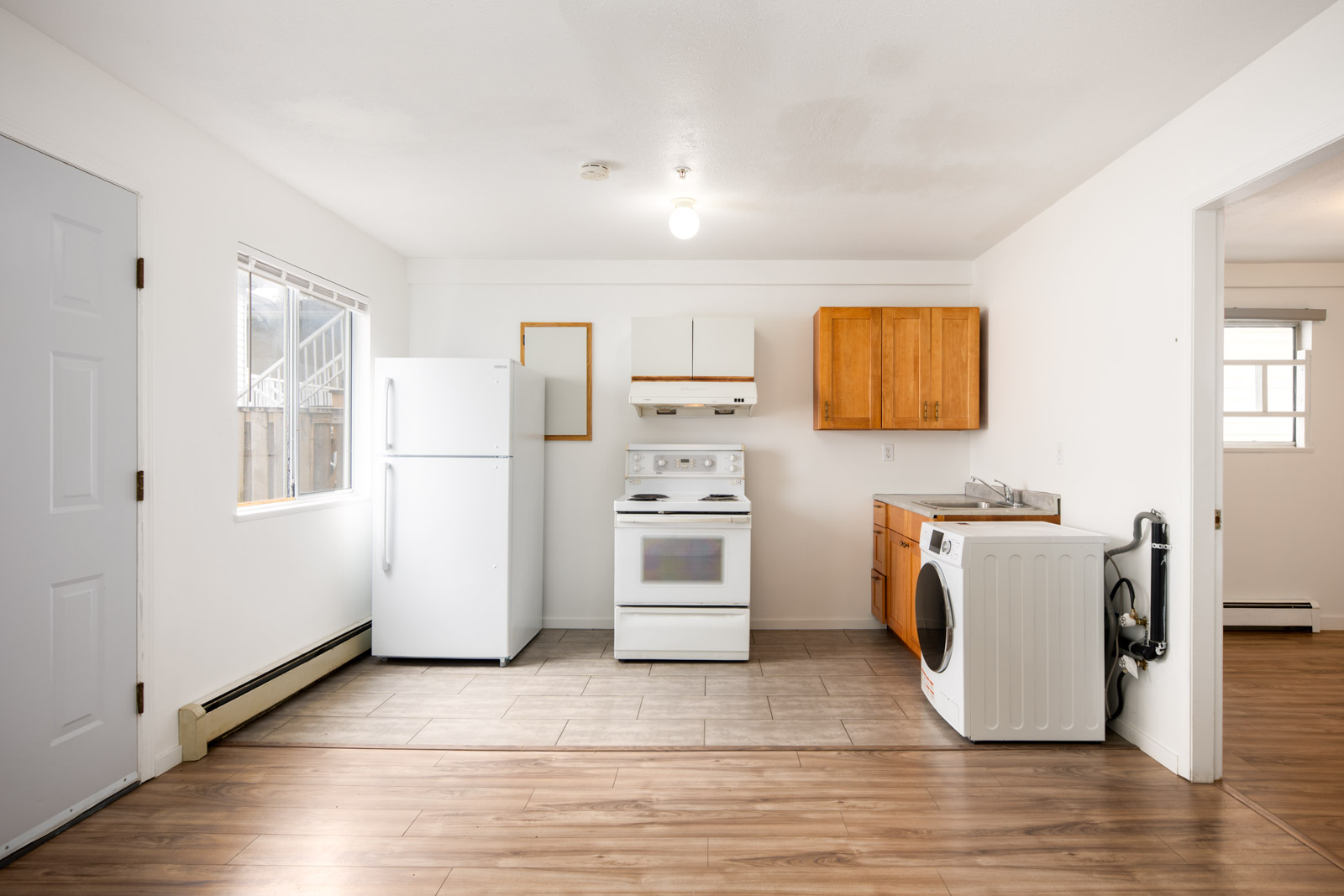 Small kitchen with white appliances, including a refrigerator, stove, and washing machine, wooden cabinets, and a window; adjacent to a bright room with wood and tile flooring.