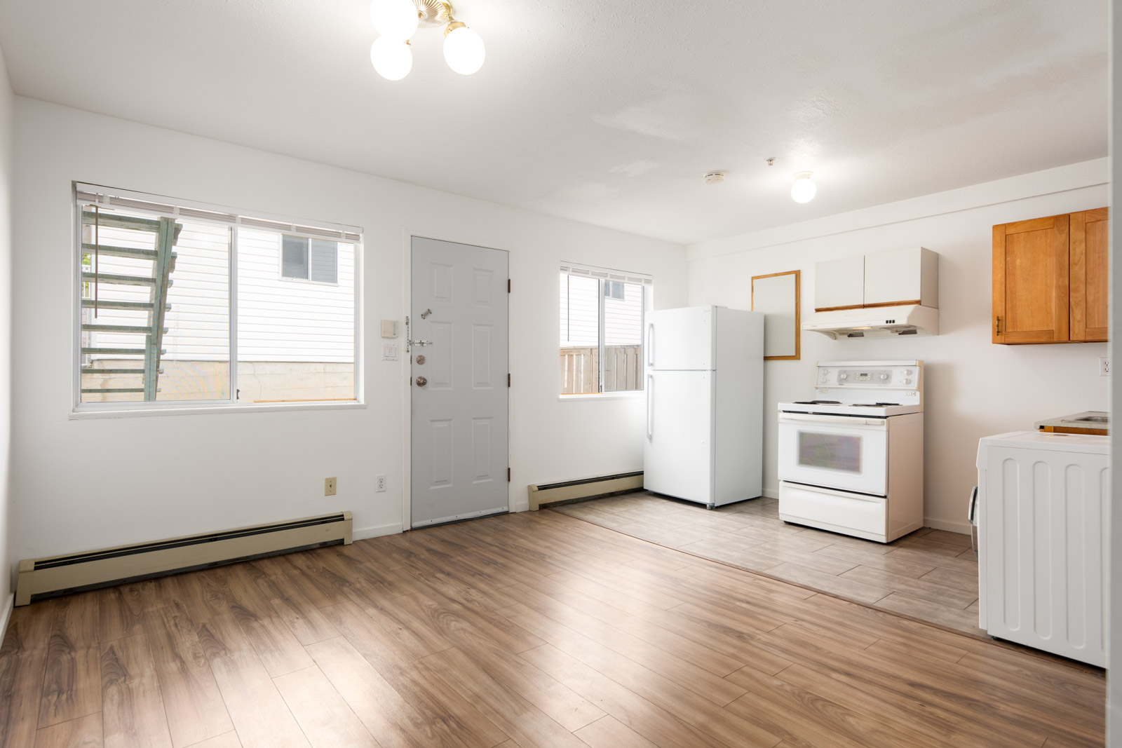 Unfurnished kitchen and living area with white walls, wood flooring, white appliances, and two large windows allowing natural light.