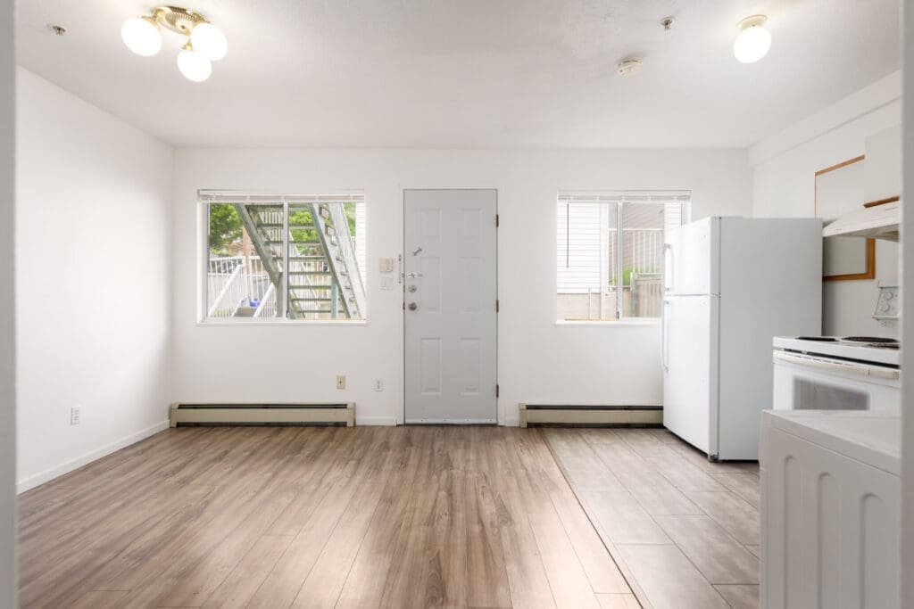 Empty apartment interior with wood-style flooring, white walls, front door, two windows, refrigerator, and stove visible; staircase is seen outside through the window.
