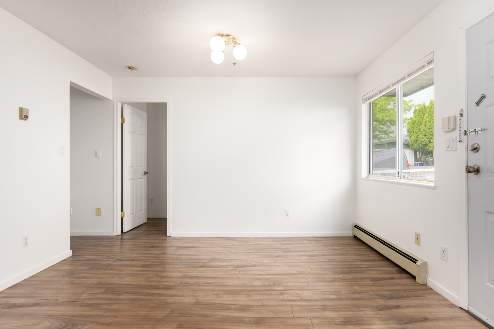 Empty room with white walls, wood flooring, a window letting in daylight, a closed white door, baseboard heater, and a ceiling light fixture.