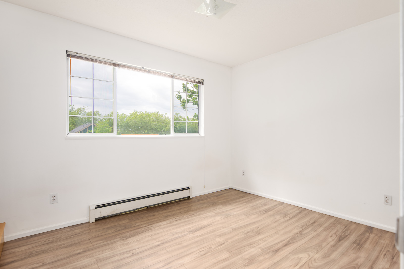 Empty room with white walls, a large window with blinds, light wood flooring, and a baseboard heater beneath the window.