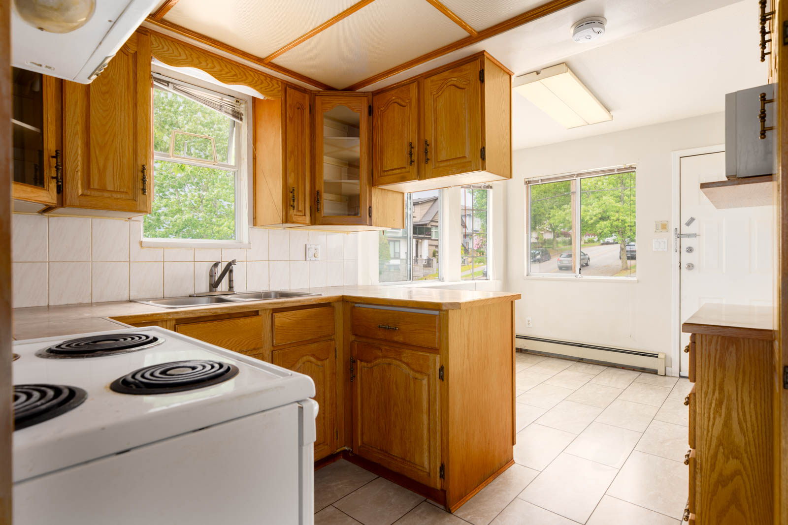 A small kitchen with wooden cabinets, white tile backsplash, electric stove, and large windows letting in natural light.