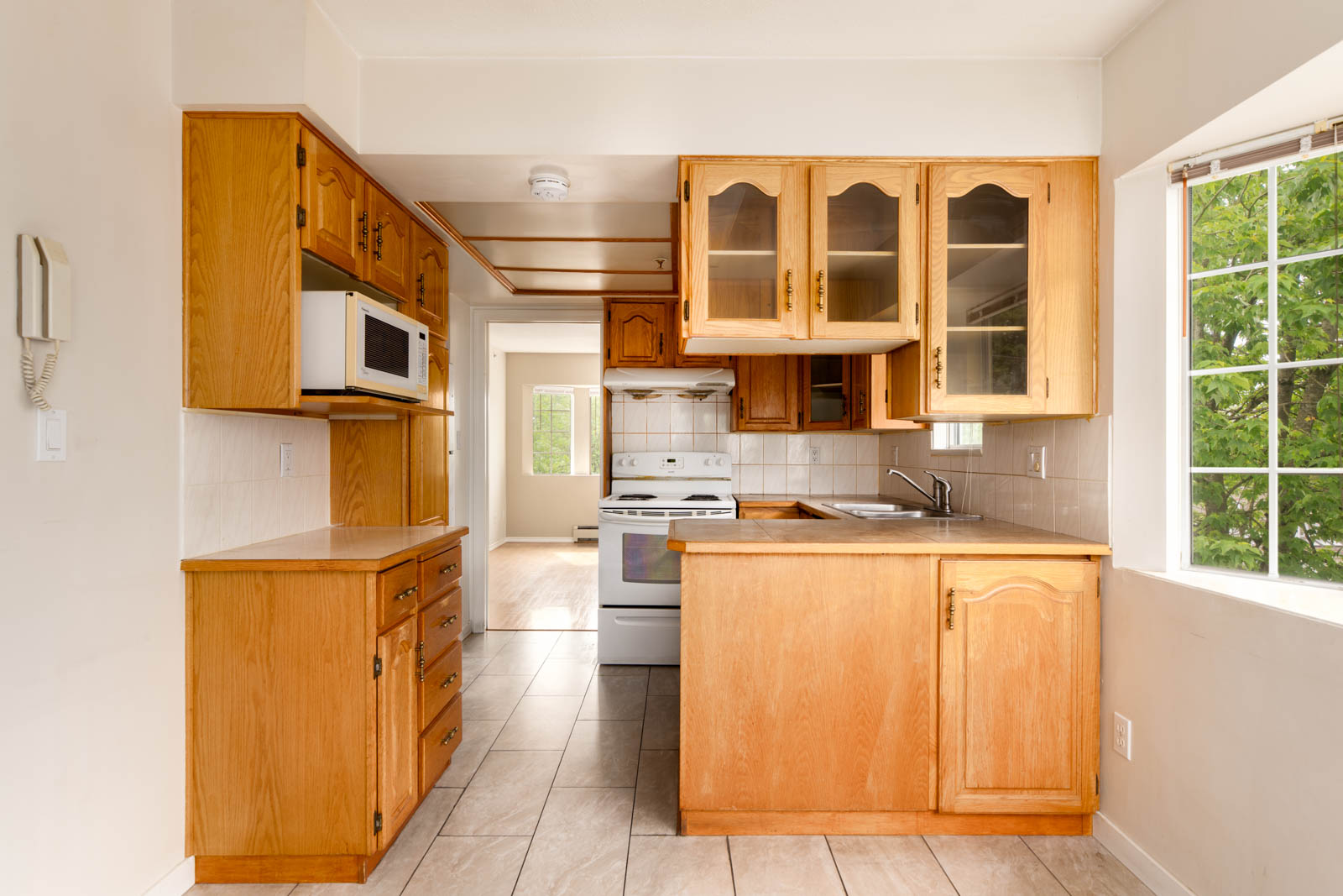 A compact kitchen with wooden cabinets, white appliances, tiled floor, and a large window letting in natural light.