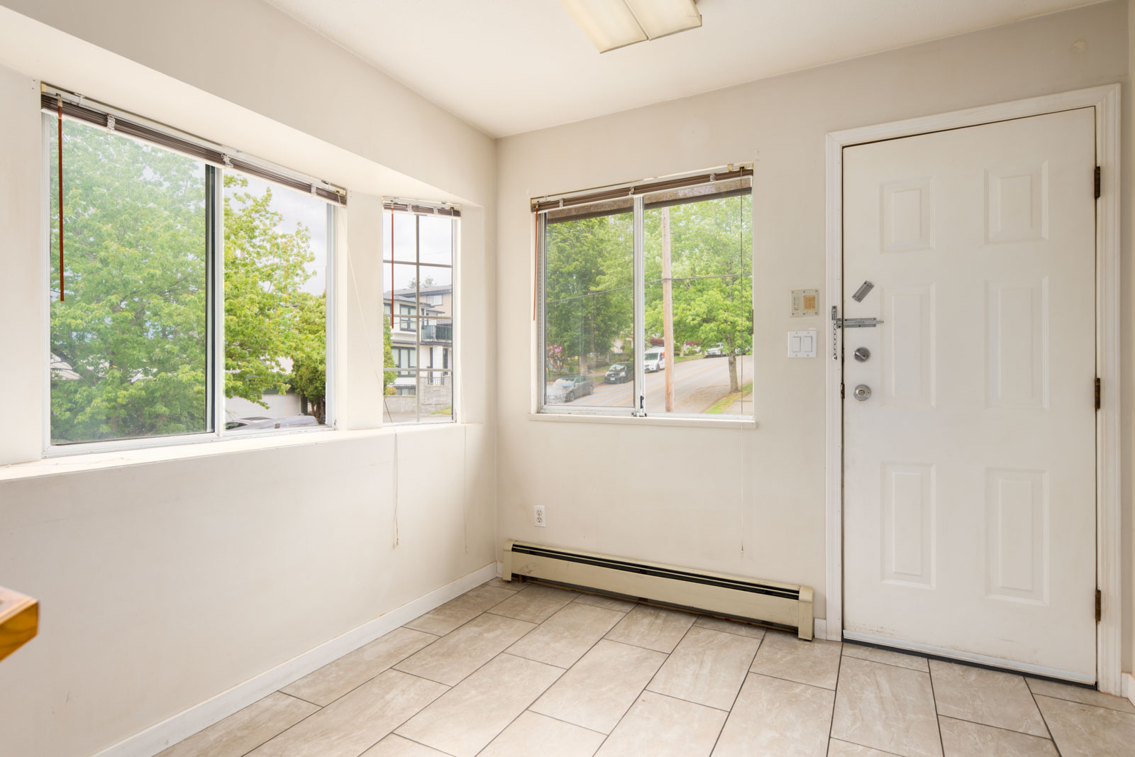 A small, empty room with tiled floor, three windows, and a white door. Trees and a street are visible outside through the windows.